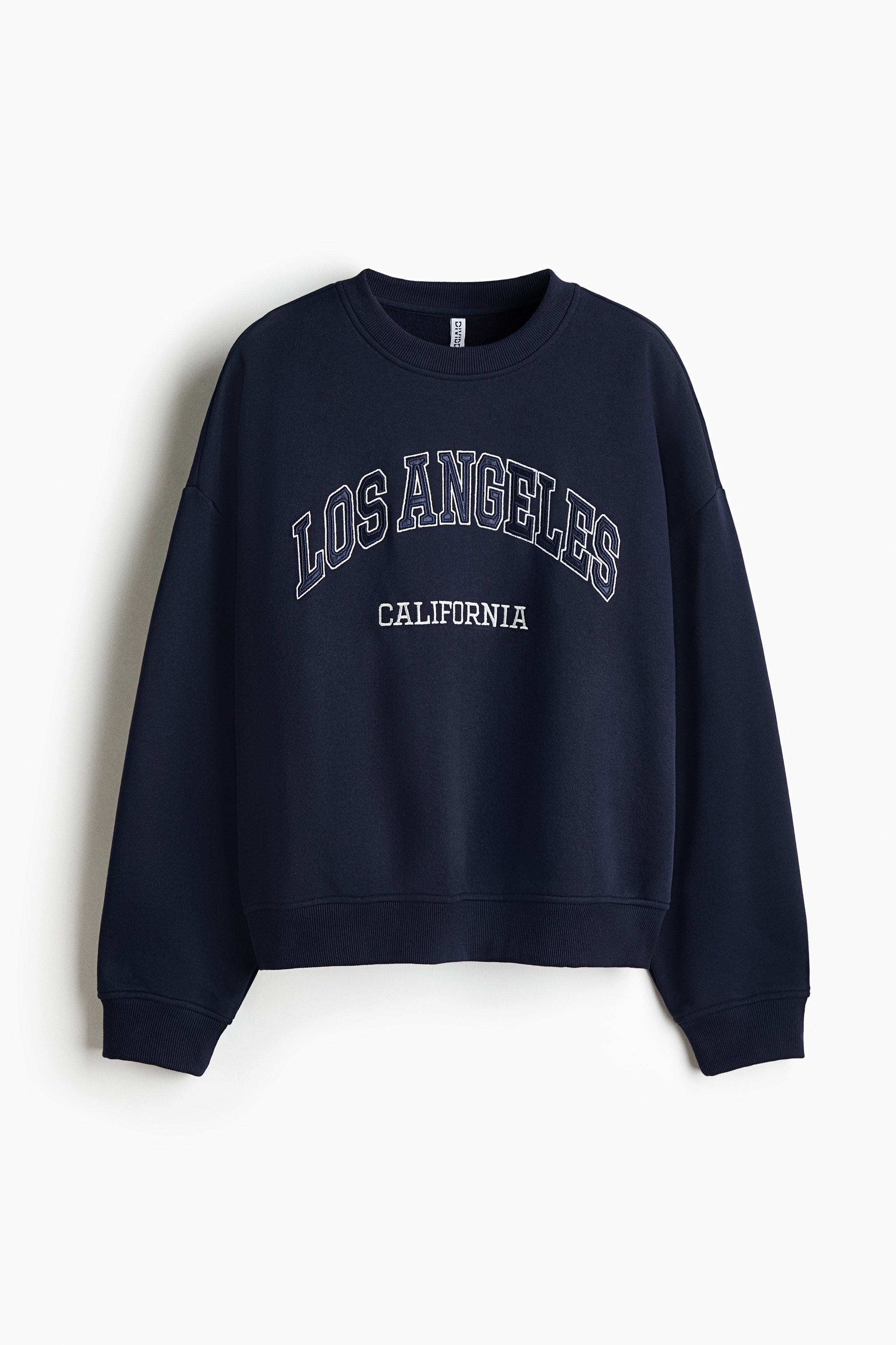 An oversized navy blue sweatshirt featuring a white "LOS ANGELES CALIFORNIA" printed design is being worn by the model over a white T-shirt. The sweatshirt has a round, rib-trimmed neckline, dropped shoulders, and ribbed cuffs, with the model raising one arm to reveal a white long-sleeve layer underneath.