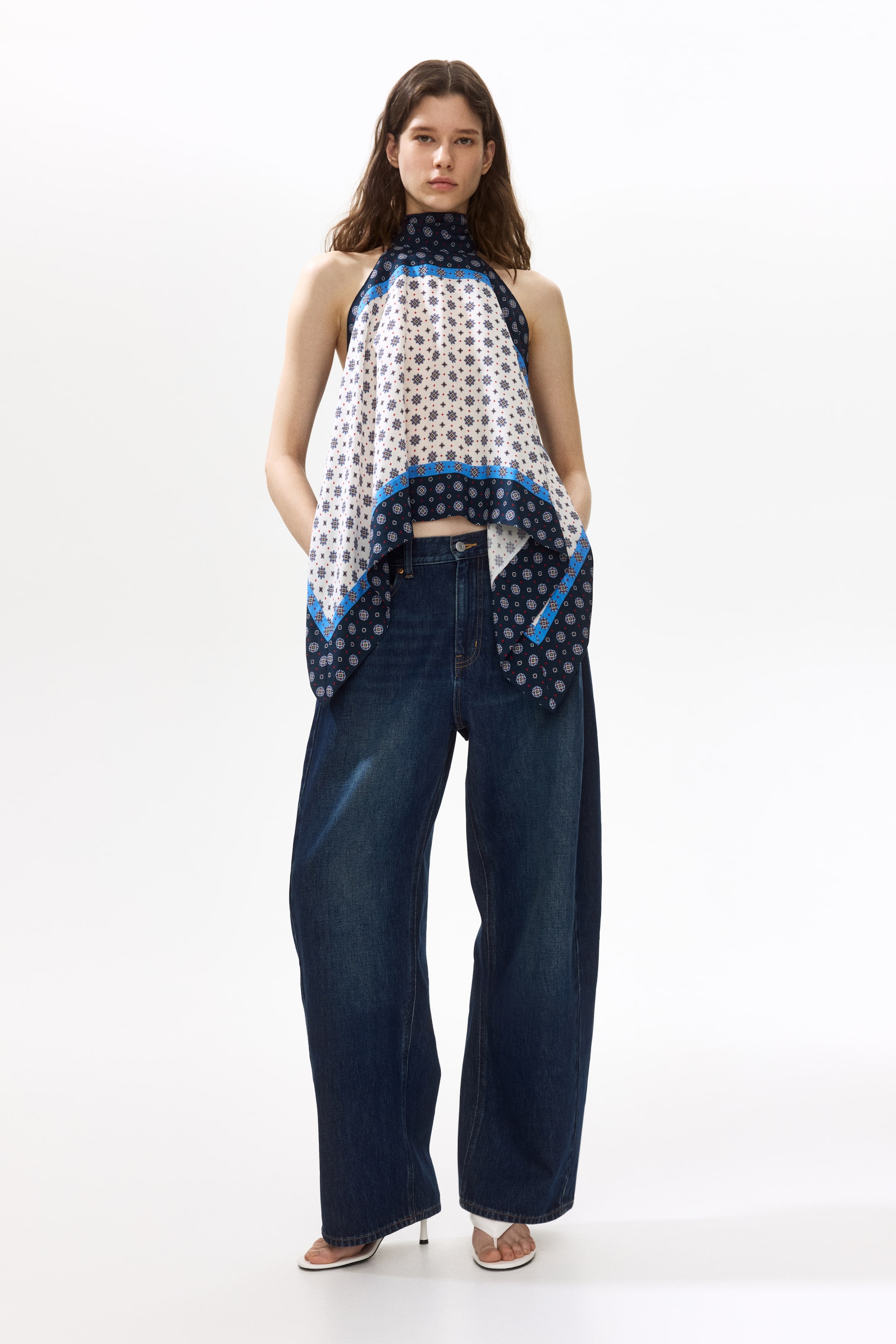 Ver imagen más grande: El producto presenta un top de cuello halter con un patrón en blanco y azul, con un escote alto y un dobladillo asimétrico en forma de pañuelo. Este top cuenta con patrones geométricos y un borde en azul oscuro que contrasta, y se combina con unos vaqueros de lavado oscuro, de talle alto y pierna ancha, que muestran un cierre frontal de botones y cremallera.