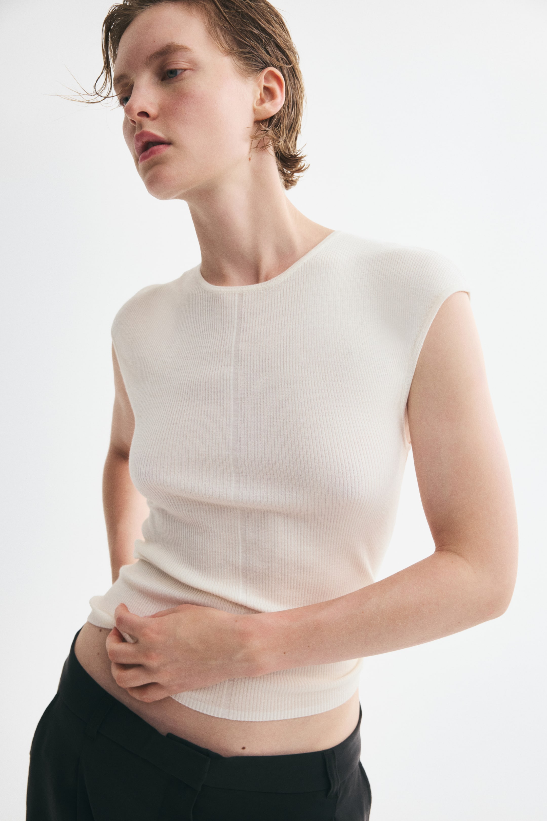 Visualizza immagine più grande: Un top corto aderente in maglia a coste, con scollo rotondo, maniche corte e orlo dritto, indossato dalla modella. La modella tiene delicatamente l'orlo del top con una mano.
