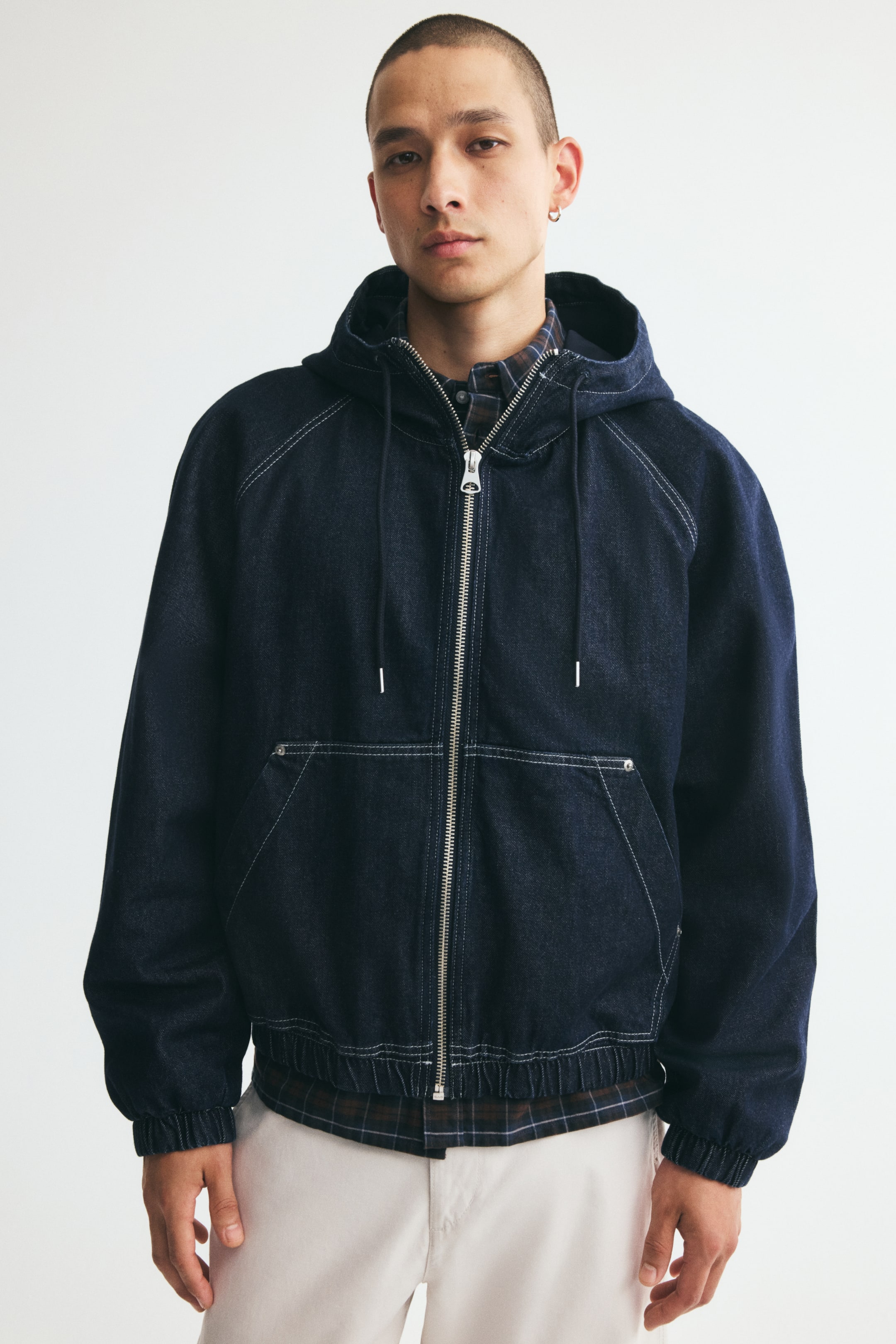 View larger image: A dark blue cotton denim jacket is being worn by the model, featuring a drawstring hood, a front zipper closure, raglan sleeves, and an elasticated hem and cuffs. The jacket, detailed with white contrast stitching and patch front pockets, is layered over a dark plaid shirt and paired with light-coloured trousers.