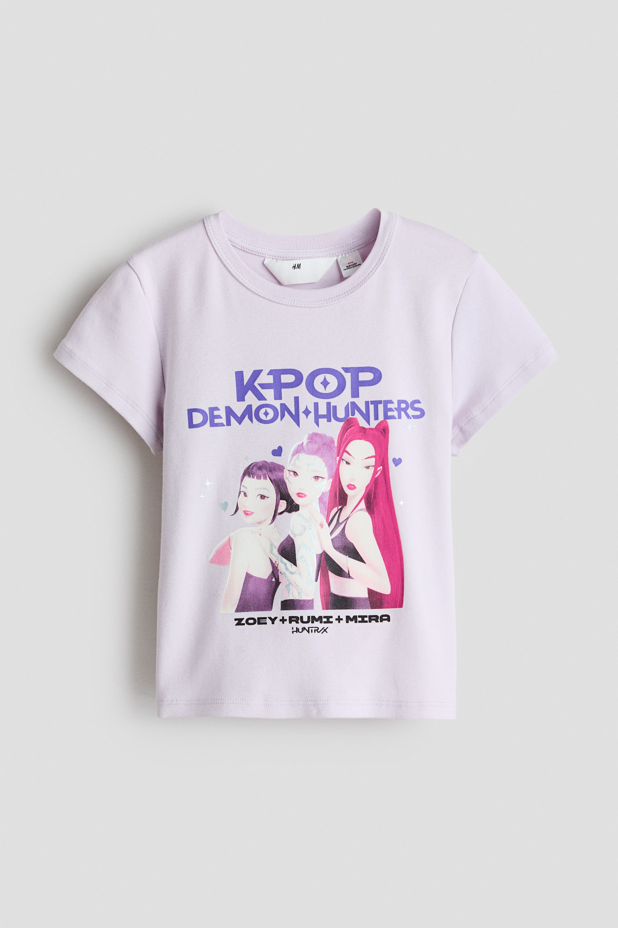 Ver imagem maior: Um top de manga curta em jersey de algodão suave, de cor roxo claro, com decote redondo. A parte frontal do top exibe um motivo impresso, incluindo o texto "KPOP DEMON HUNTERS" acima de três personagens femininas estilizadas e "ZOEY + RUMI + MIRA HUNTRX" abaixo delas.