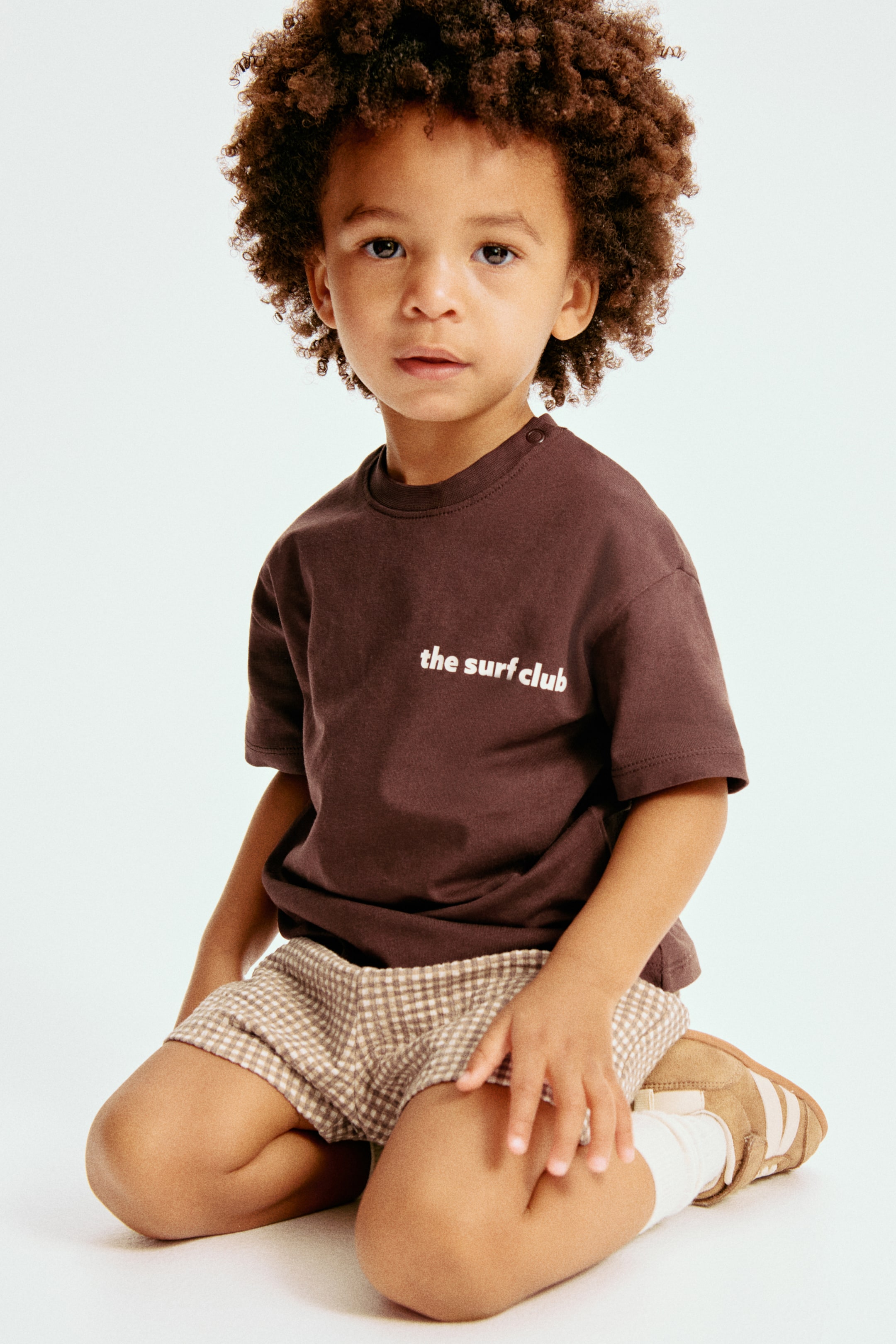 View larger image: A dark brown short-sleeved t-shirt with white "The Surf Club" text on the chest and snap buttons on the left shoulder is paired with light brown and white checkered shorts, cream ankle socks, and tan shoes featuring white stripes.
