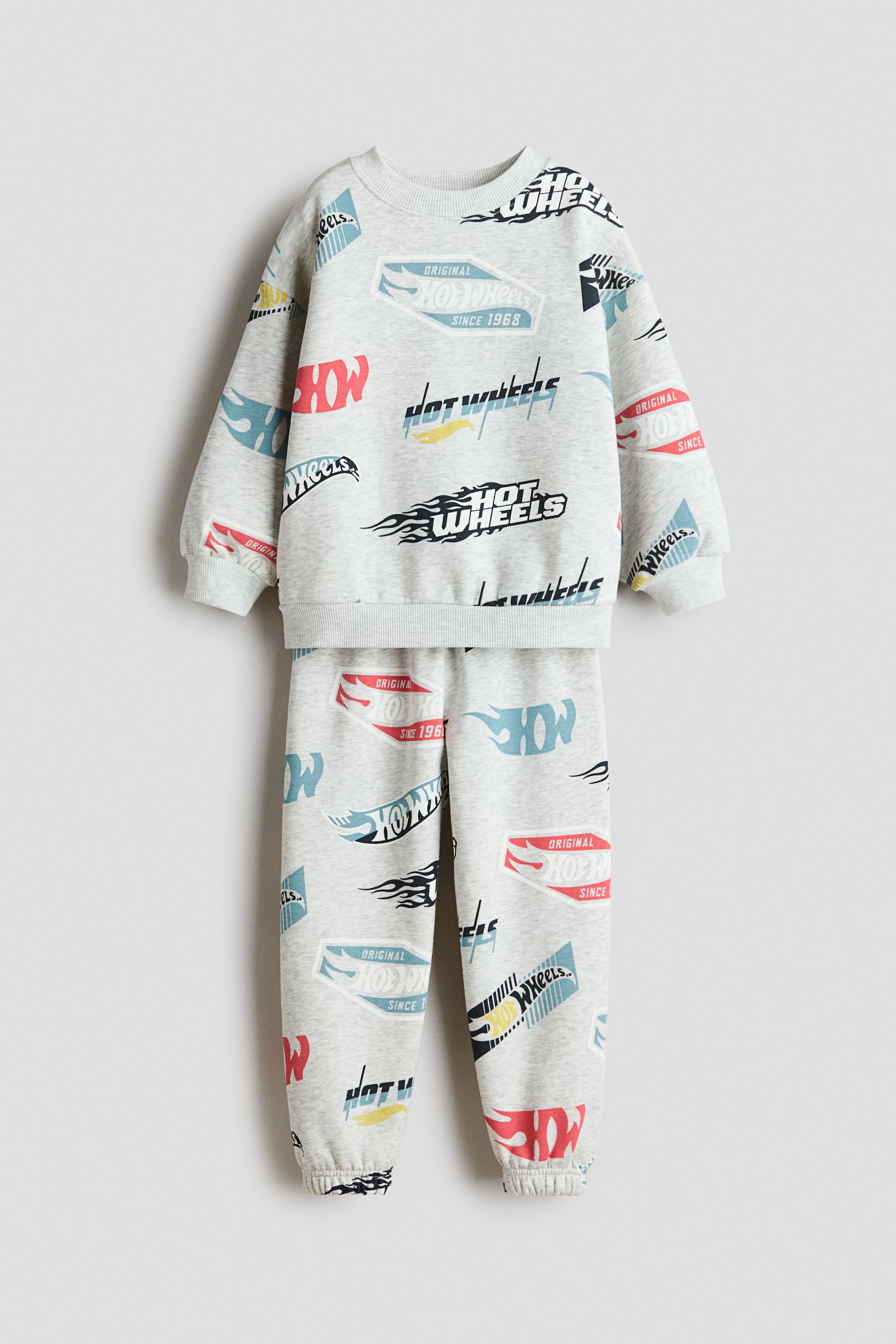 Grotere afbeelding bekijken: Een lichtgrijze Hot Wheels loungewear set wordt FRONT weergegeven, bestaande uit een sweatshirt met ronde hals en bijpassende joggingbroek. Beide stukken hebben een all-over print van kleurrijke Hot Wheels-logo's en tekst; het sweatshirt is ontworpen met geribde boorden en een geribde zoom, en de joggingbroek is afgewerkt met elastische enkels.