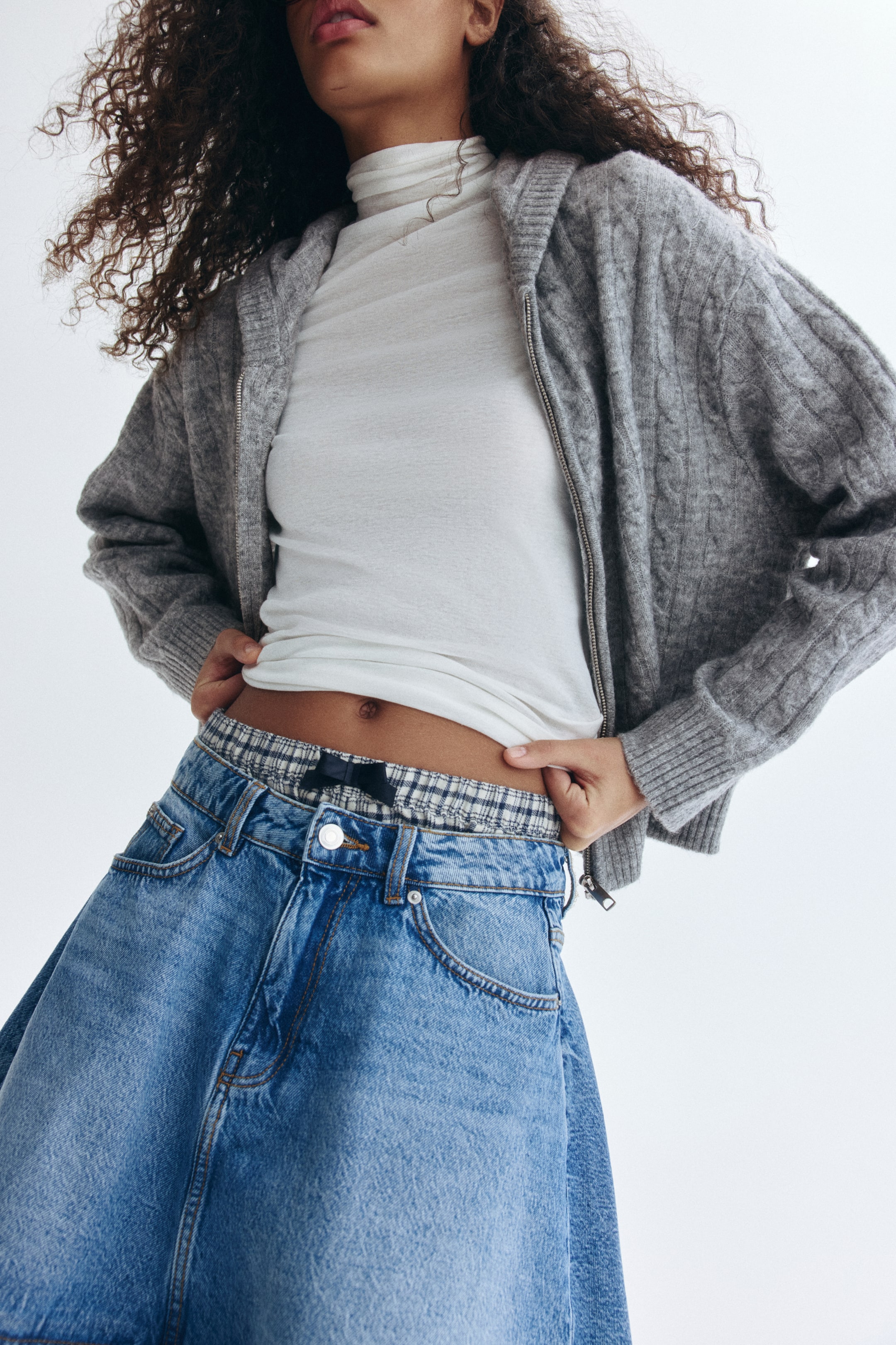 Ingrandisci l'immagine: Un cardigan con cappuccio a maglia intrecciata di colore grigio, con una chiusura anteriore con cerniera e polsini a coste, è indossato. La modella tiene l'orlo inferiore del cardigan, tirandolo leggermente verso l'alto, mentre è stratificato su un top a collo alto bianco.