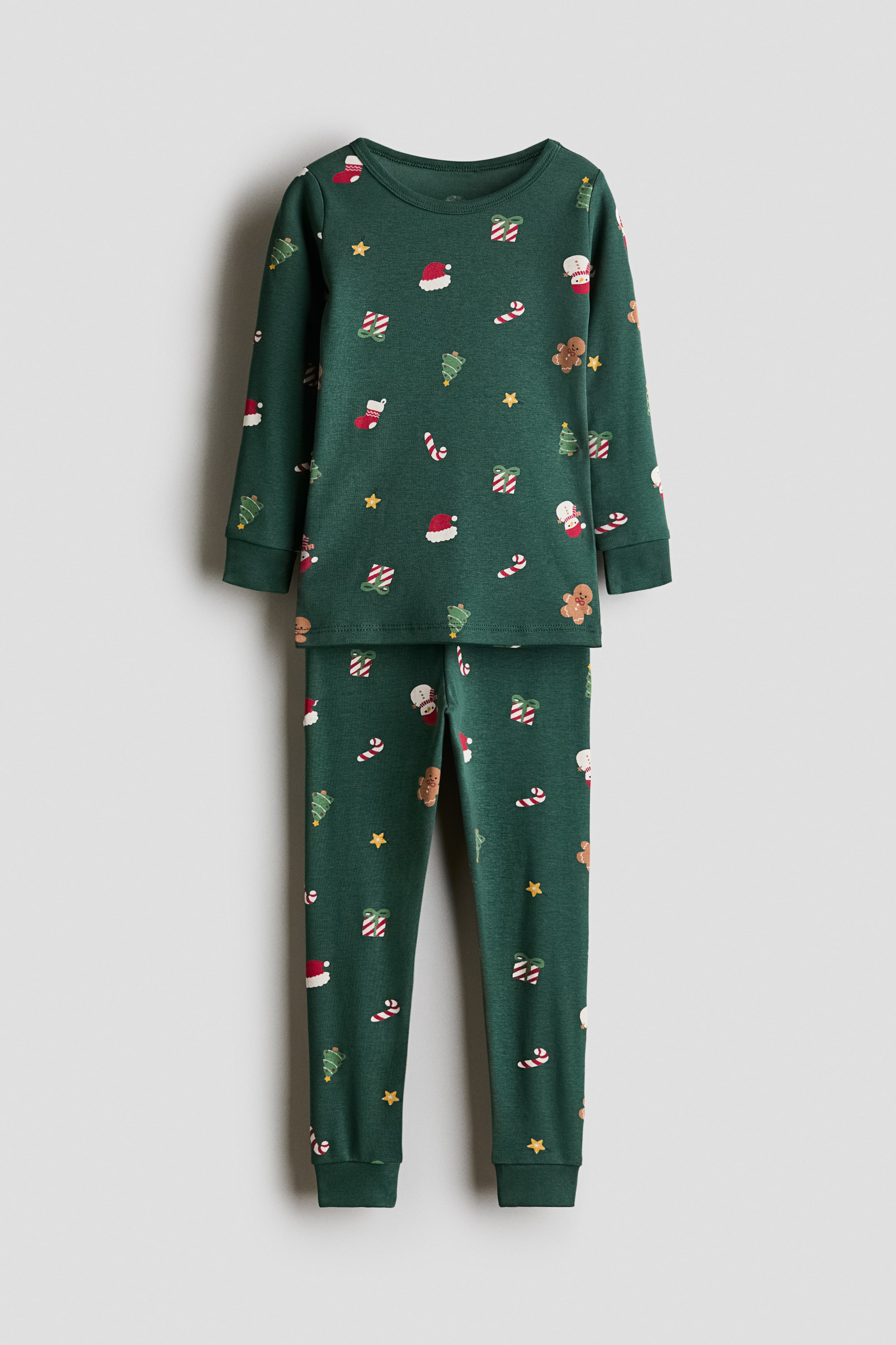 View larger image: A dark green cotton jersey pajama set is shown from the front, featuring an all-over festive print of gingerbread men, candy canes, and holiday trees. The top has a round neckline and long, cuffed sleeves, while the matching pants feature an elasticated waist and cuffed hems.