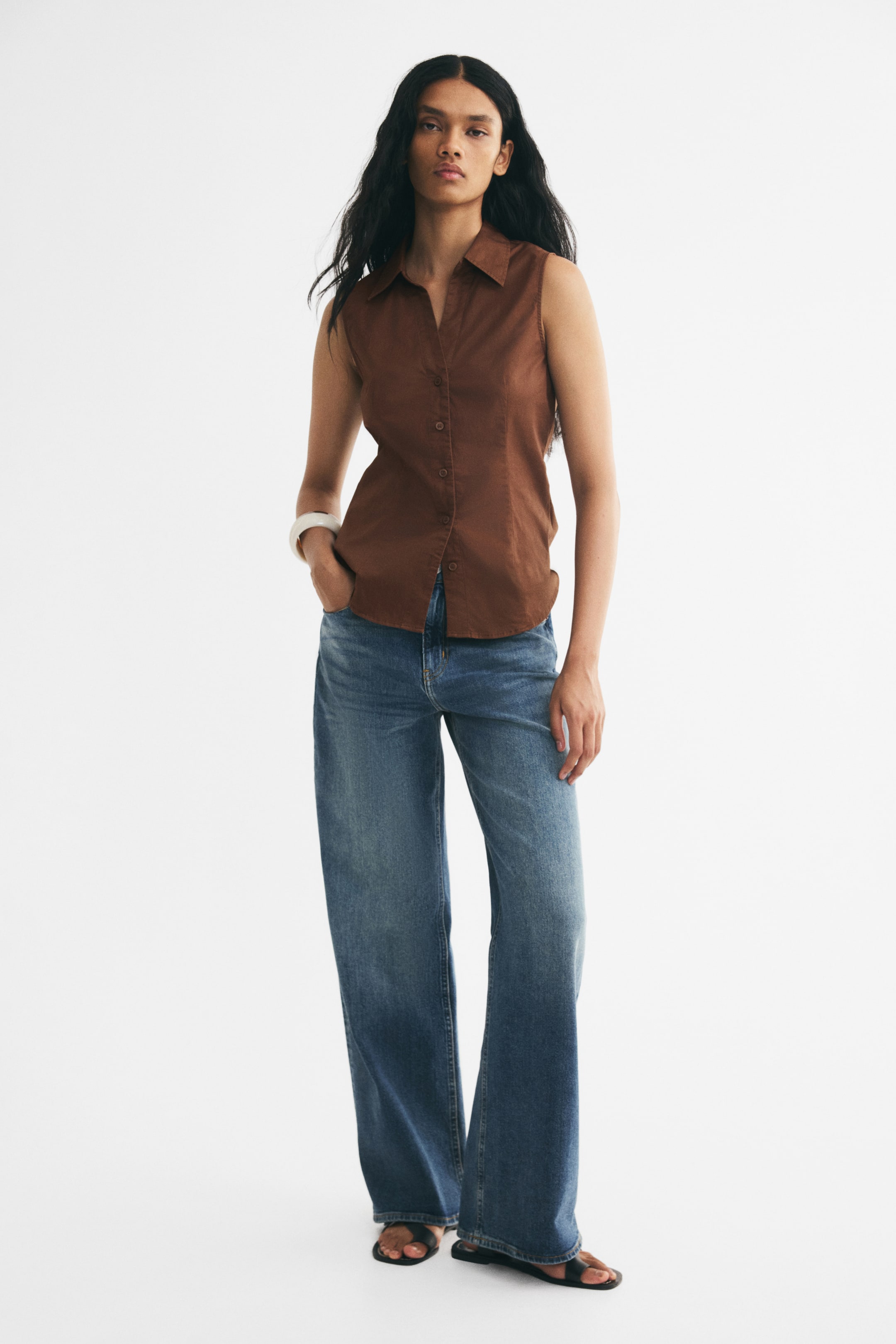 View larger image: A sleeveless, fitted brown shirt with an open collar, front buttons, and a rounded hem is worn by the model. The shirt is styled with wide-leg blue jeans, with the model standing and placing one hand in a front pocket.