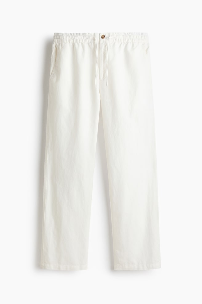White trousers are shown from the front, featuring a gathered elasticated waistband with a central drawstring tie and a light brown button, side pockets, and a straight, regular leg silhouette. - 1
