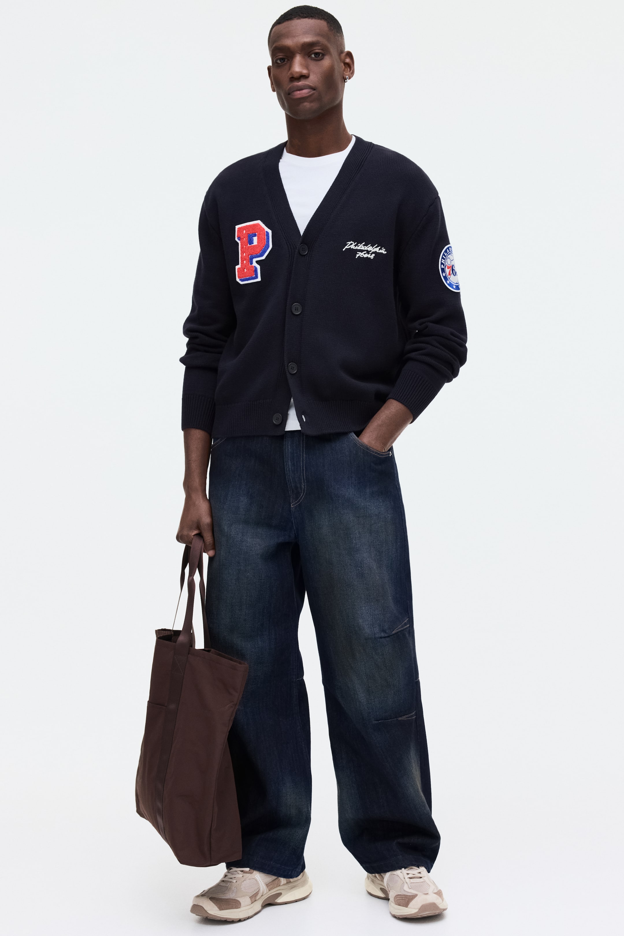 View larger image: A dark blue V-neck cardigan features a button-front closure, ribbed cuffs, and a ribbed hem, worn over a white t-shirt. It displays a large red and blue 'P' appliqué on the left chest, white embroidered 'Philadelphia 76ers' text, and a circular patch on the right sleeve.
