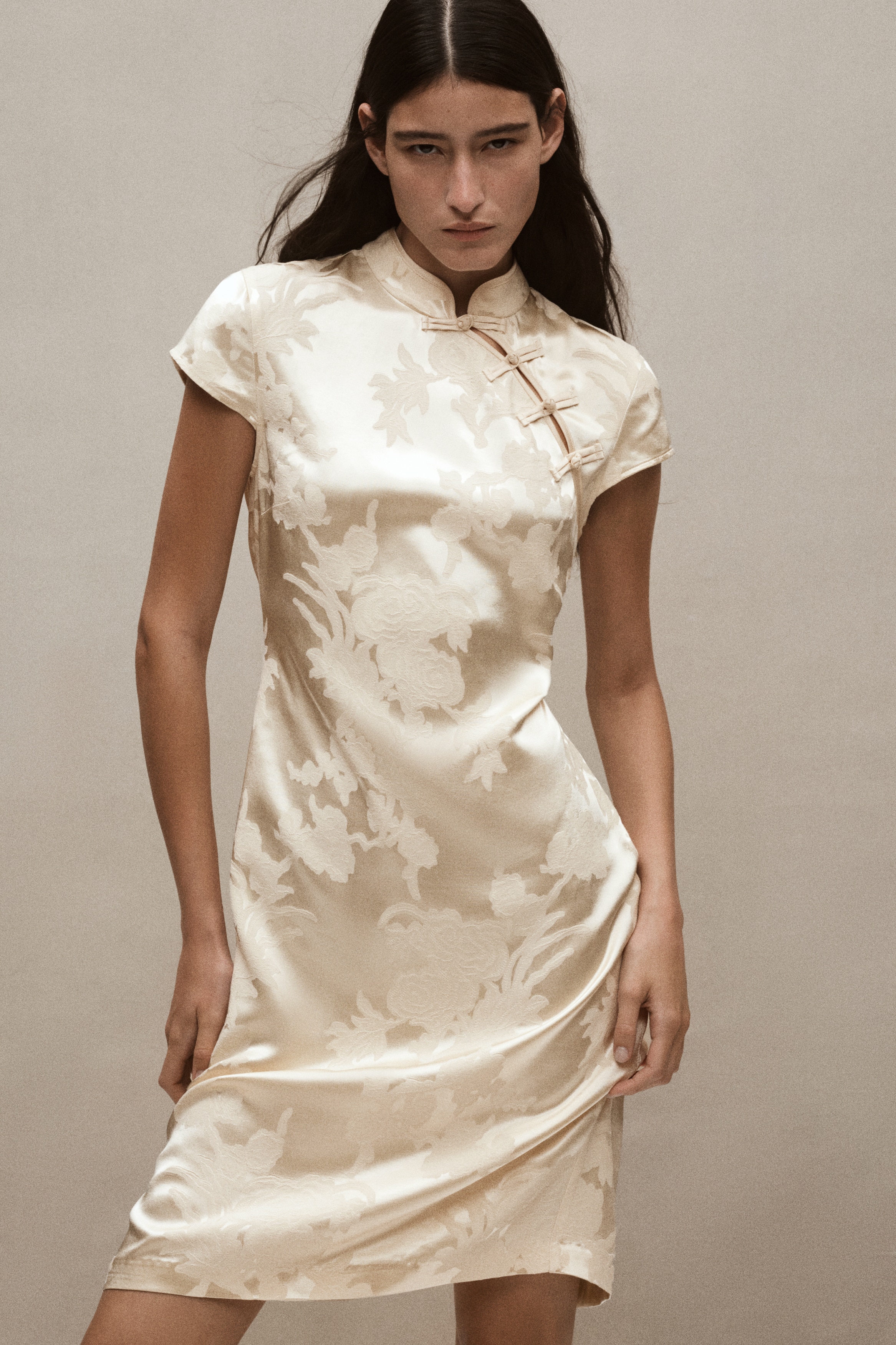 A cream-coloured, knee-length fitted dress in a floral jacquard weave is hanging facing front, showcasing its subtle sheen. It features a stand collar, short cap sleeves, and a diagonal slit opening at the neckline secured with three knot buttons.