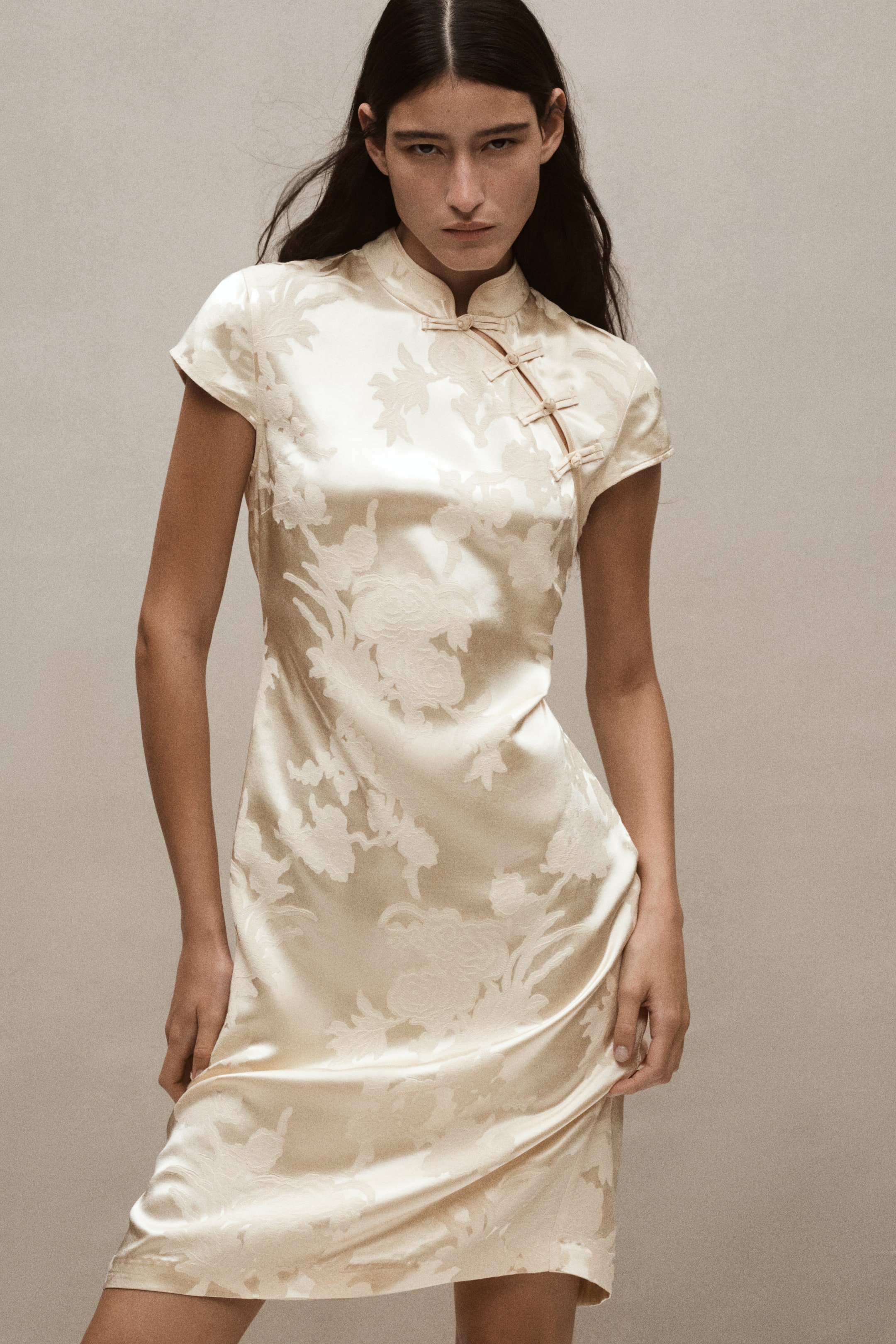 Ampliar la imagen: La modelo lleva puesto un vestido entallado color crema, a la altura de la rodilla, en tejido jacquard con acabado brillante. El vestido presenta cuello mao, mangas cortas y una abertura diagonal con botones tipo nudo en la parte superior.