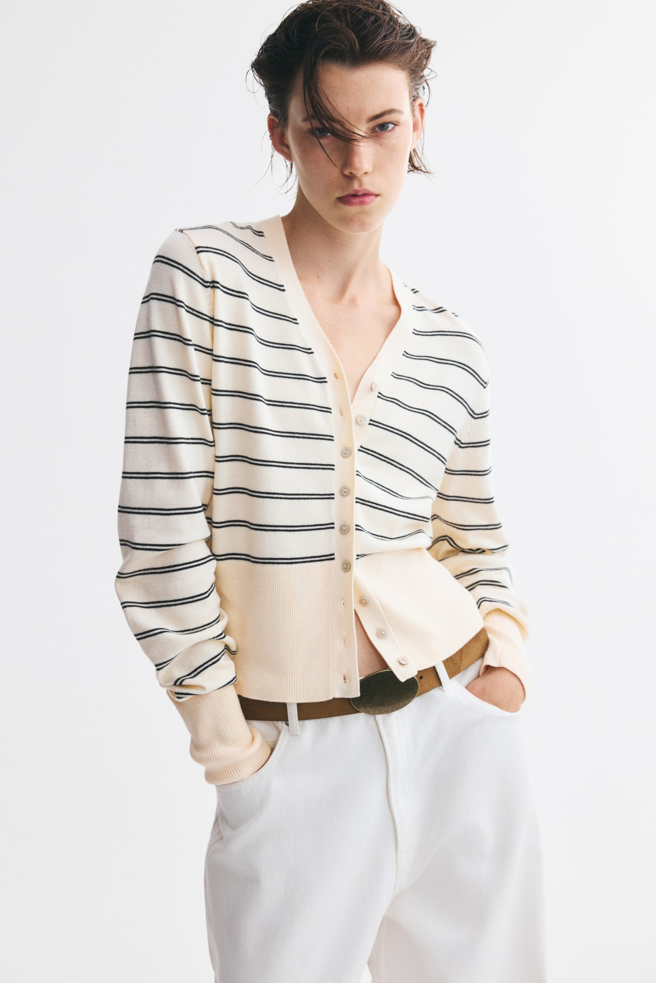 Agrandir l'image: Un cardigan court et ajusté couleur crème à fines rayures horizontales noires, porté de face, présente un col en V, une fermeture boutonnée sur le devant et des manches longues avec de larges poignets côtelés unis couleur crème. Le cardigan est montré partiellement déboutonné à l’ourlet large côtelé uni couleur crème, laissant apparaître une ceinture marron et un pantalon blanc.