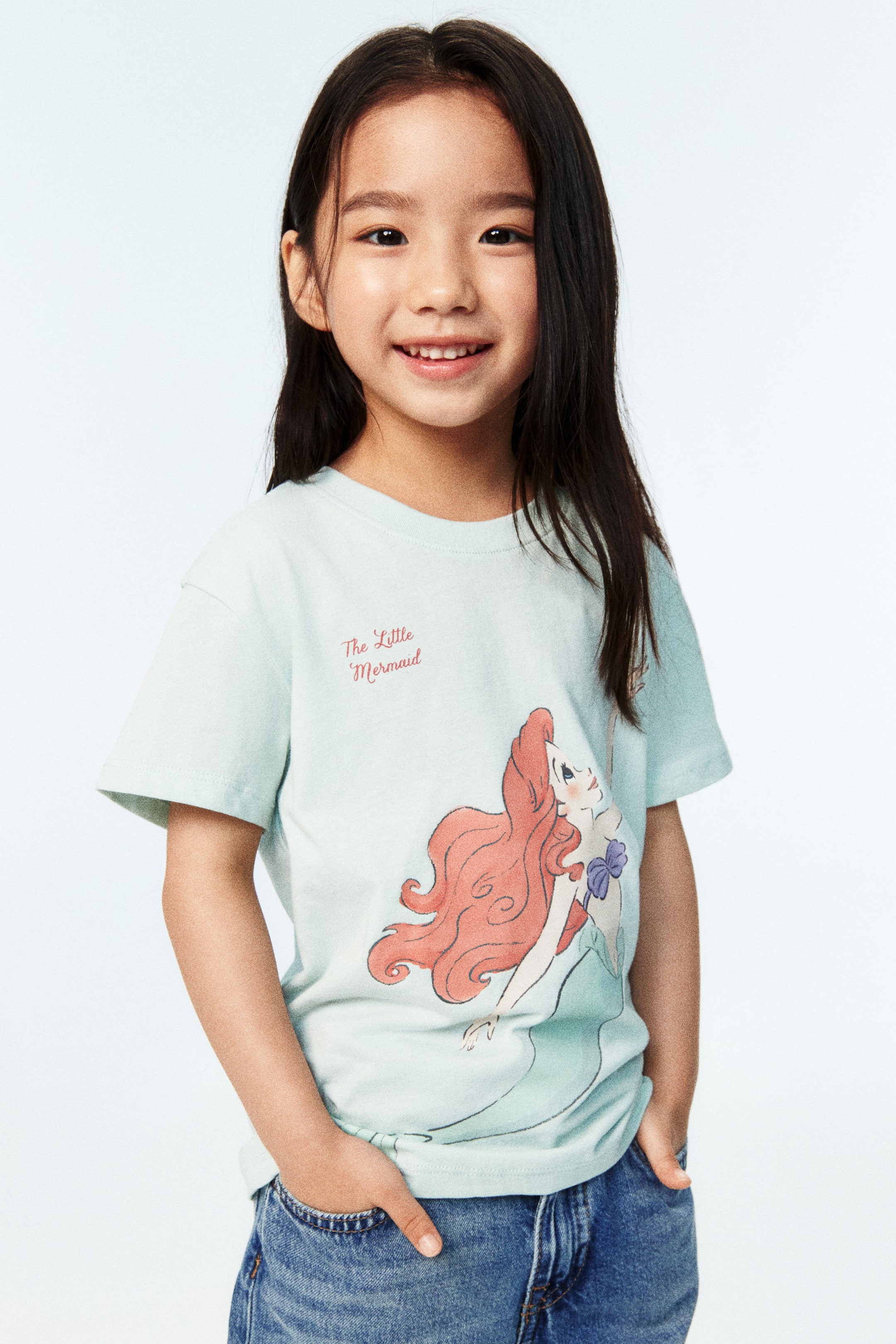 View larger image: A light green T-shirt in soft cotton jersey, featuring a print motif of "The Little Mermaid" text and an illustration of Ariel on the front, is being worn by the model. This T-shirt is designed with a round, rib-trimmed neckline and gently dropped shoulders, and is being styled with blue denim jeans.