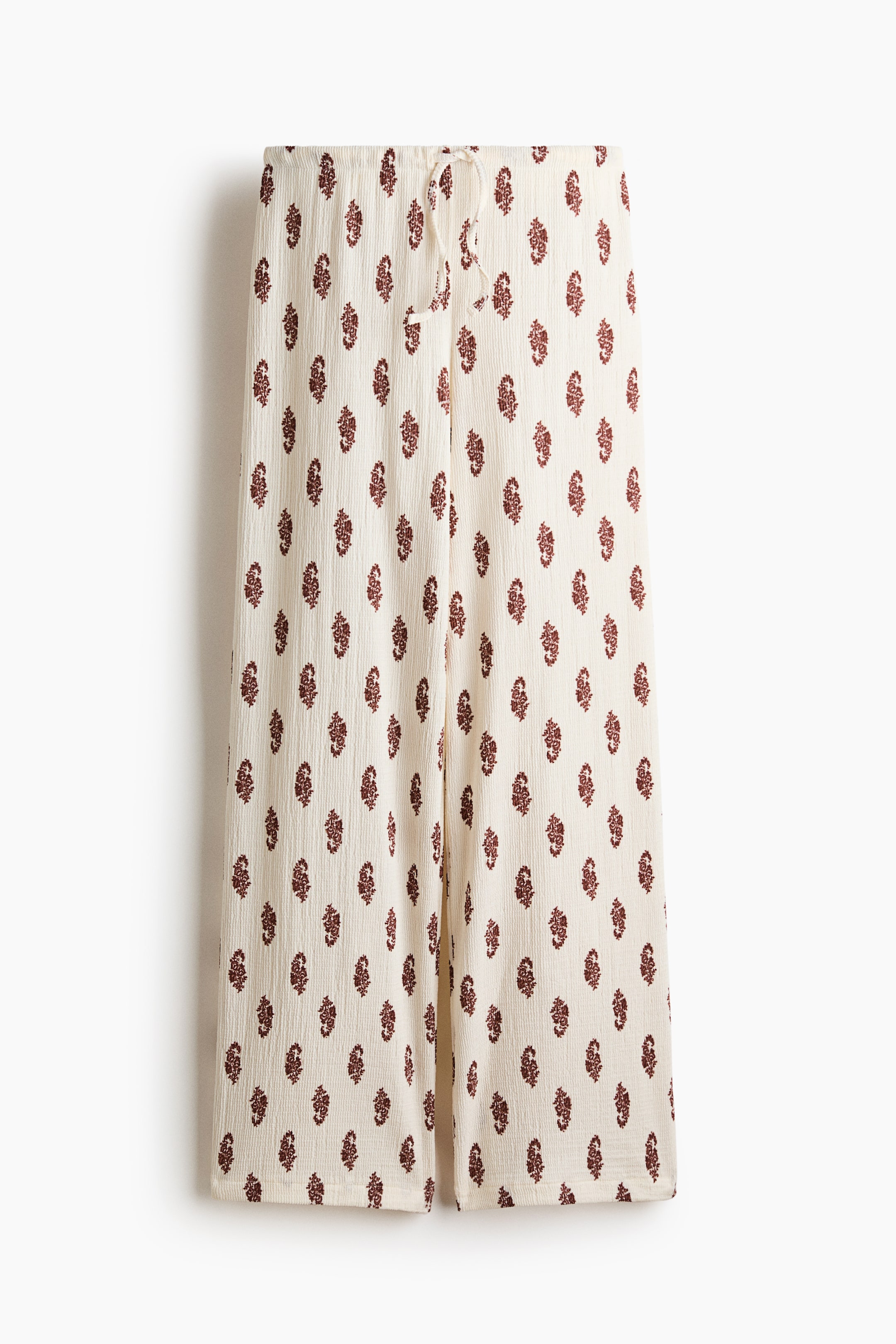 View larger image: The trousers showcase a front view, crafted from a textured cream jersey fabric adorned with repeating maroon paisley-like motifs. At the top, there's an elasticated, drawstring waist, while straight legs extend downwards.
