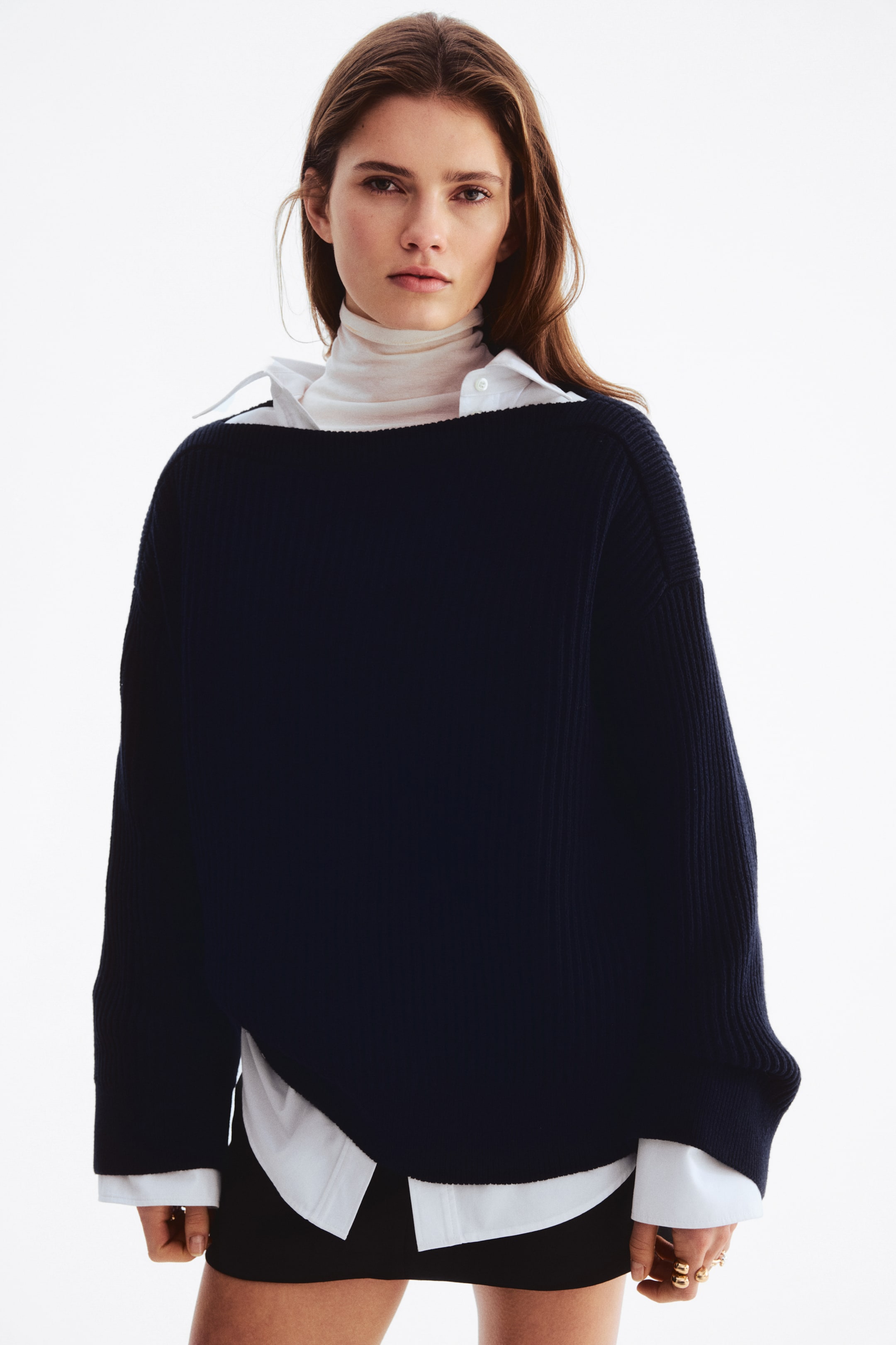 View larger image: A navy blue oversized sweater, featuring a wide boat neck, vertical ribbed detailing on the sleeves and side panels, and a high-low hem with side slits, is layered over a white collared shirt and a light-colored turtleneck.