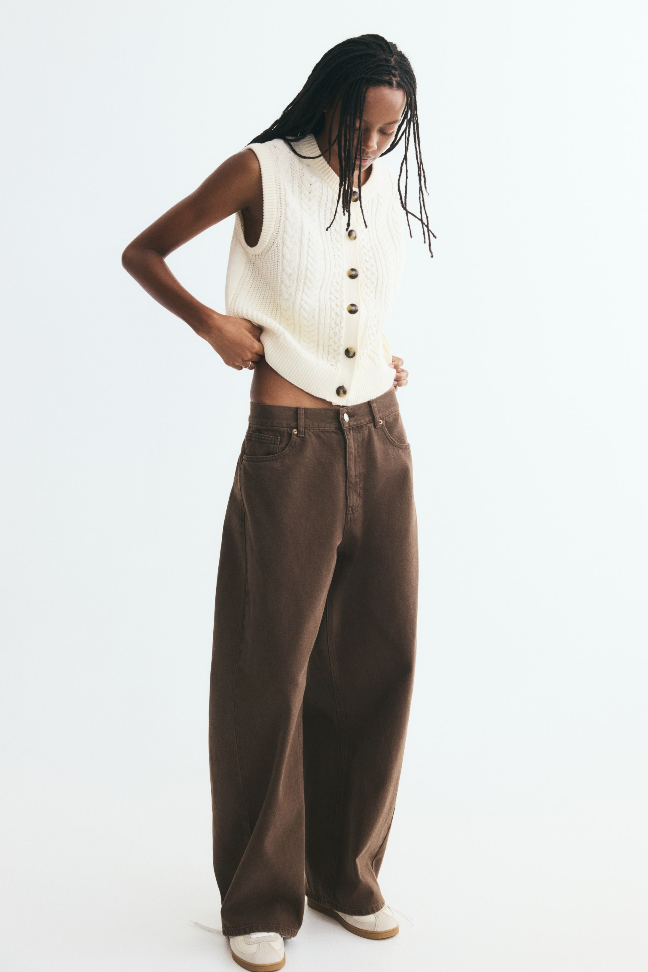 View larger image: A short, cream-coloured knit vest is shown from the front, featuring a cable knit pattern, a round ribbed neckline, and dark buttons down the front. The sleeveless vest is displaying ribbed trim along the armholes and hem, and is styled with wide-leg brown trousers.