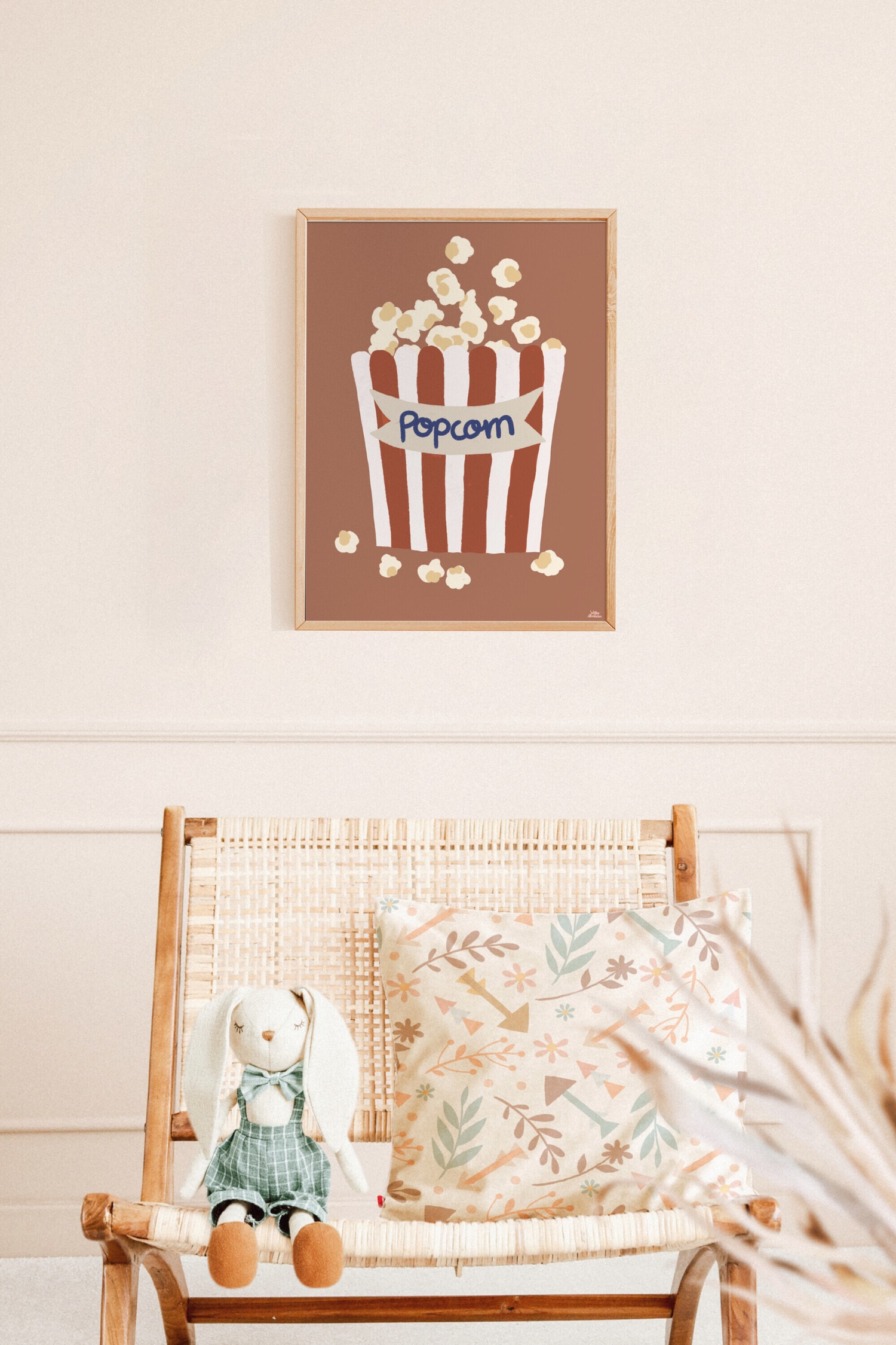 Een kunstprint toont een gestileerde illustratie van een popcornbakje met verticale bruine en witte strepen tegen een effen terracottakleurige achtergrond. Een lichtgrijze banner aan de voorkant toont het woord 'Popcorn' in blauw schrift, terwijl er meerdere crÃ¨megekleurde korrels over de rand vallen en rond de basis verspreid liggen.