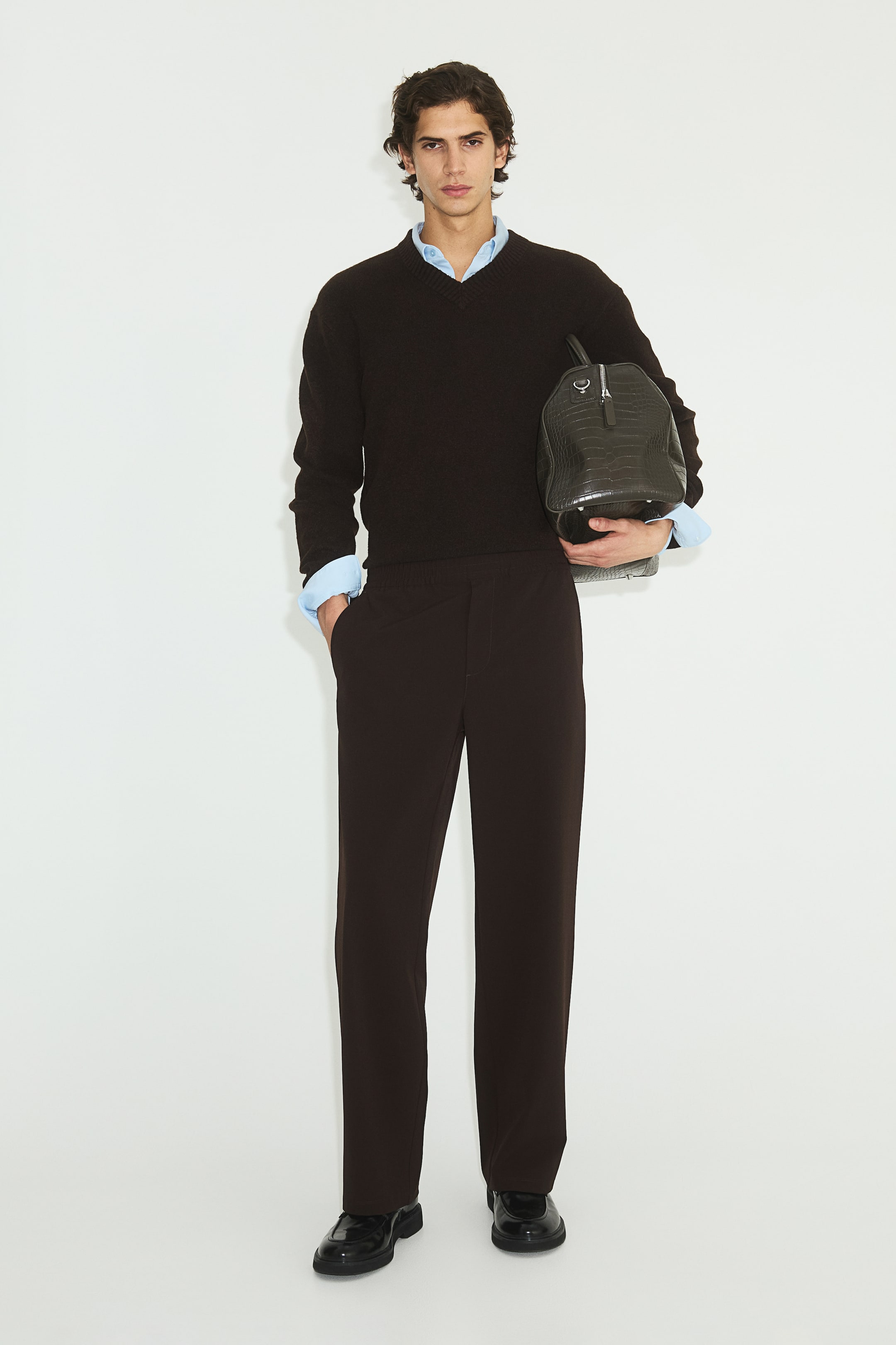 Se større billede: En mørkebrun V-hals sweater med afslappet pasform, lange ærmer med ribbede manchetter og ribbet kant, er lagdelt over en lyseblå skjorte med krave, hvor skjortens krave og manchetter er synlige.