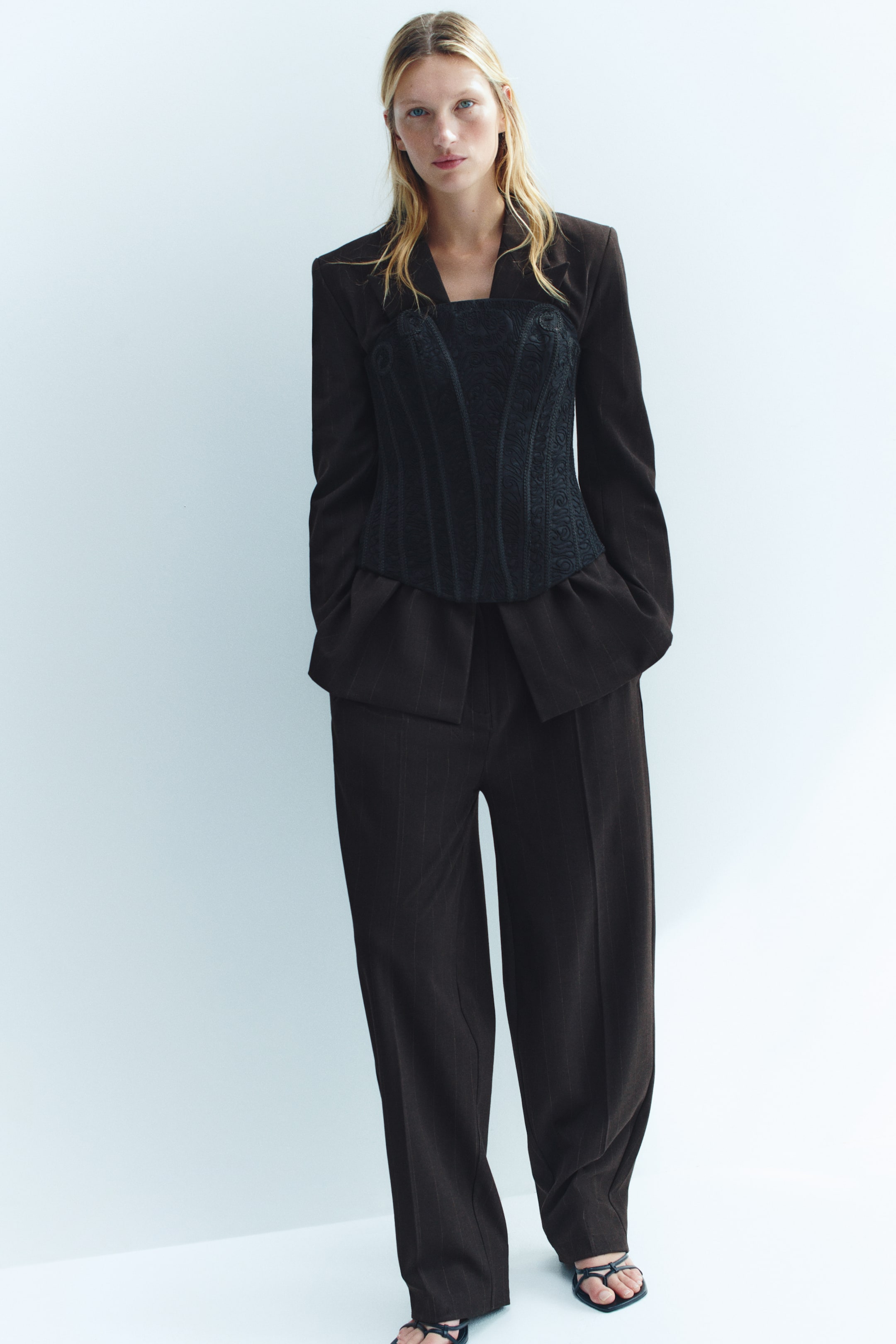 Grotere afbeelding bekijken: De model draagt een op maat gemaakte pantalon met krijtstreep, met een wijde pijpen silhouet en zichtbare vouwen in het midden. De pantalon is gestyled met een bijpassende blazer, een zwarte gestructureerde corsetlaag over het jasje en zwarte sandalen met bandjes.