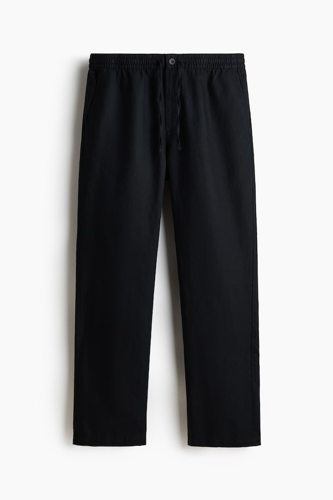 Black trousers are lying flat, presenting a front view, and featuring a covered elasticated waist with a drawstring and a single button closure. Two side pockets are visible, and the trousers are designed with a straight-leg cut. - 1