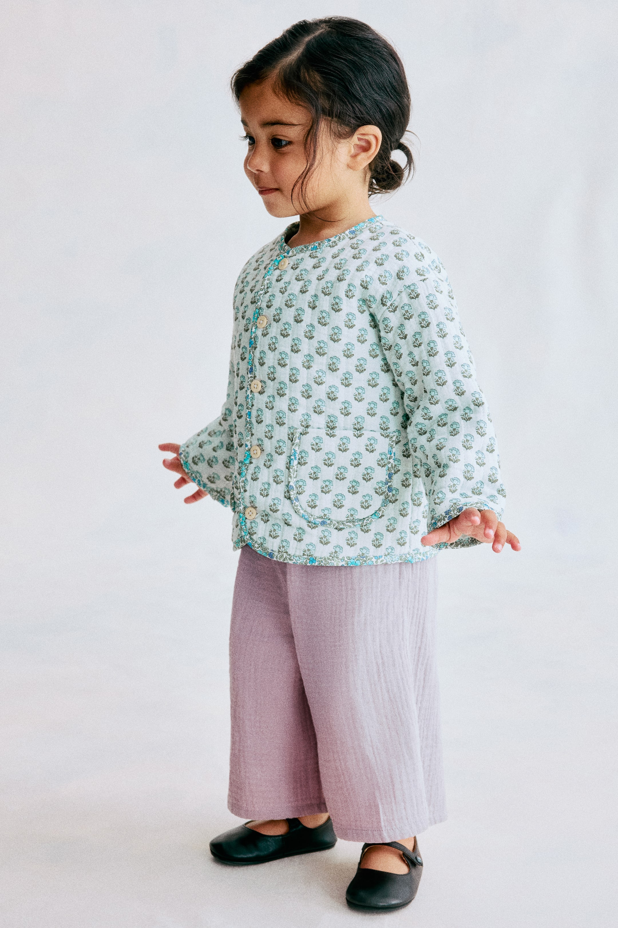 View larger image: An H&M Adorables jacket, viewed from a slight right angle, highlights a light blue quilted weave adorned with a repeating patchwork pattern. It features a round neckline, button-down front, dropped shoulders, and contrasting blue bindings along the neckline, front opening, hem, and two open front pockets.