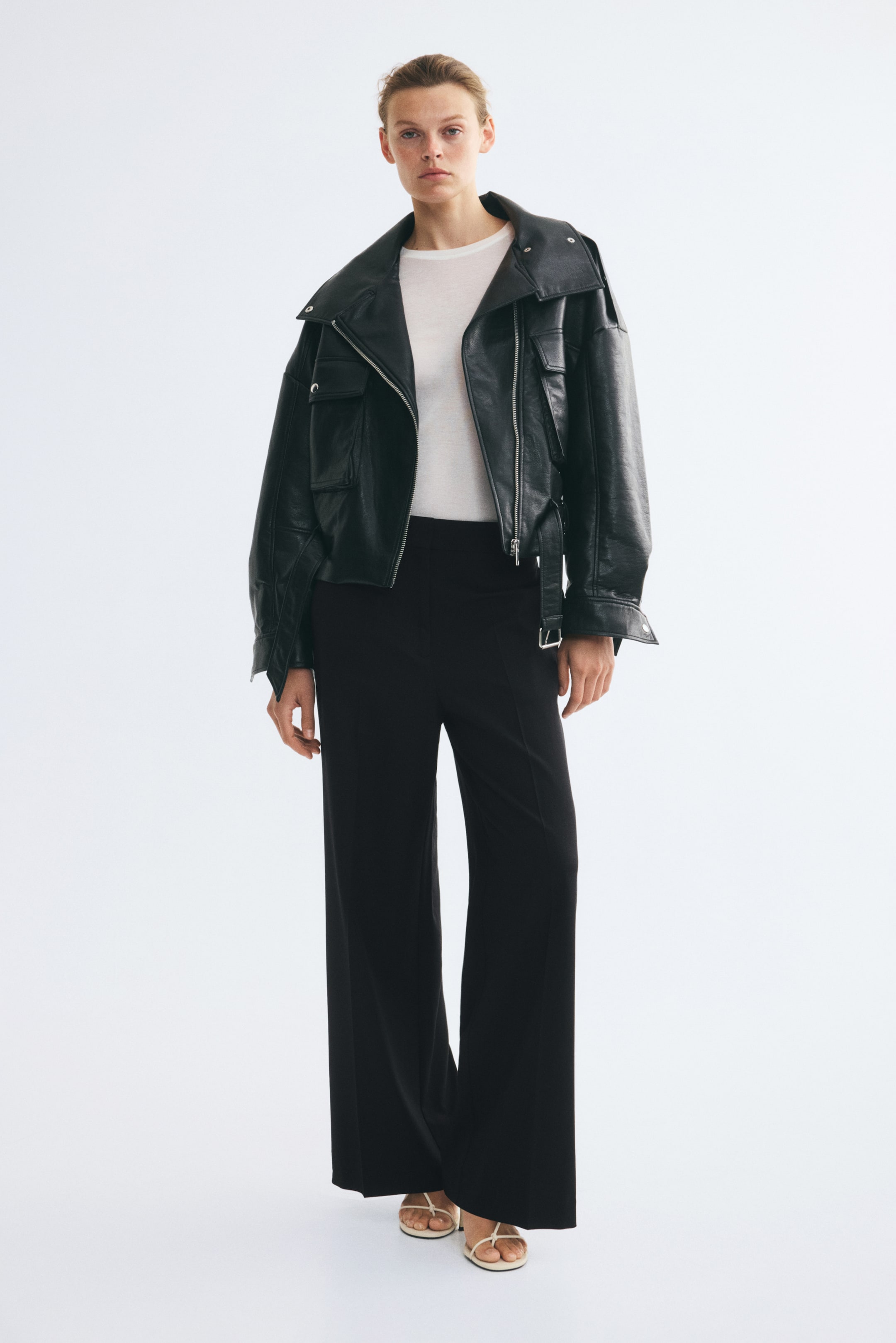 Agrandir l'image: Le mannequin porte un pantalon noir ajusté à taille haute, à jambes larges, avec des plis marqués sur le devant. Le pantalon est associé à un haut blanc rentré, à une veste oversize en cuir noir et à des sandales blanches à brides.