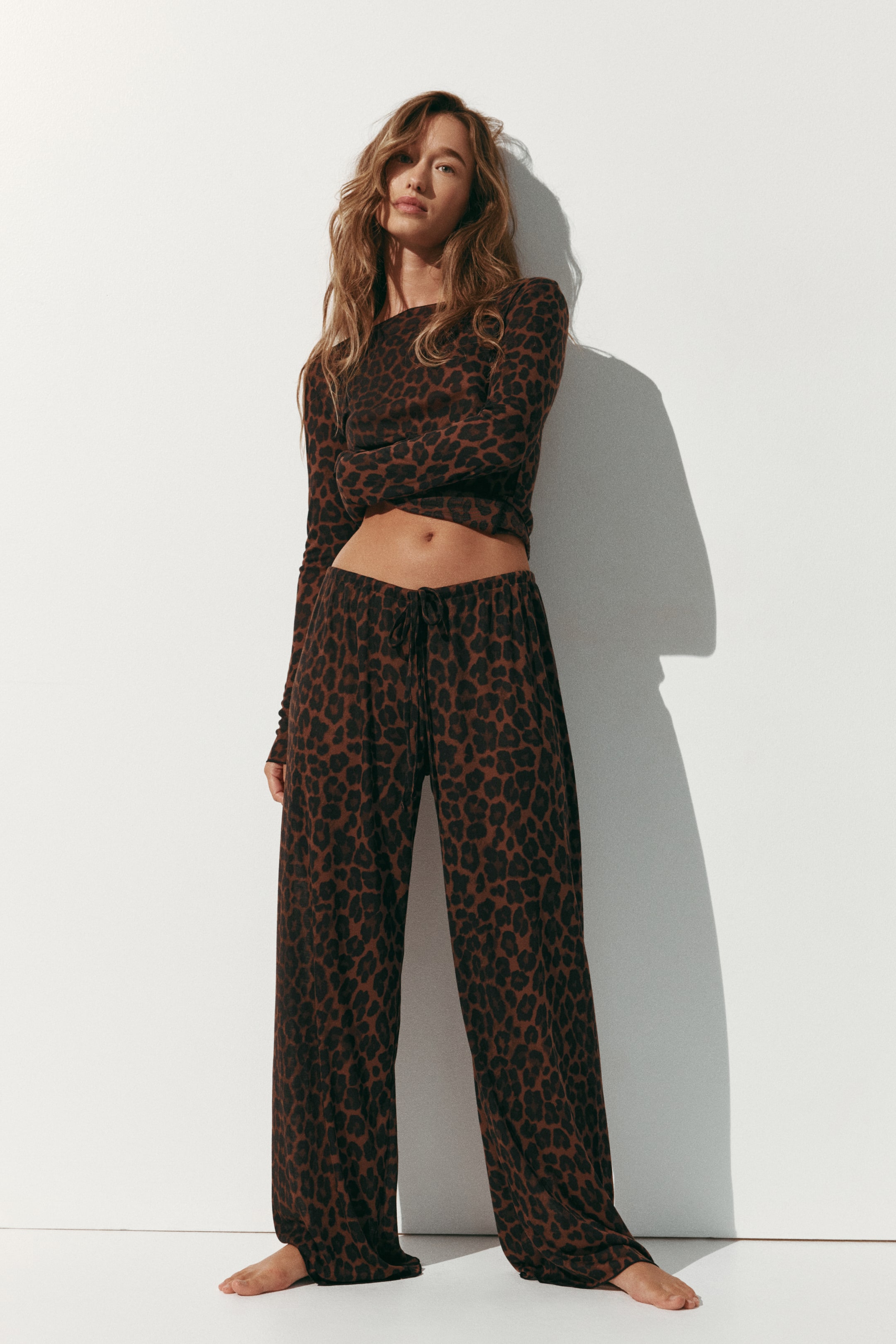 View larger image: A leopard-print pyjama set is shown from the front, featuring a fitted, long-sleeved top with a boat neckline and matching wide-leg pants. The pants display an elasticised, drawstring waist, and both pieces showcase a sheer, ribbed jersey fabric with overlocked edges.