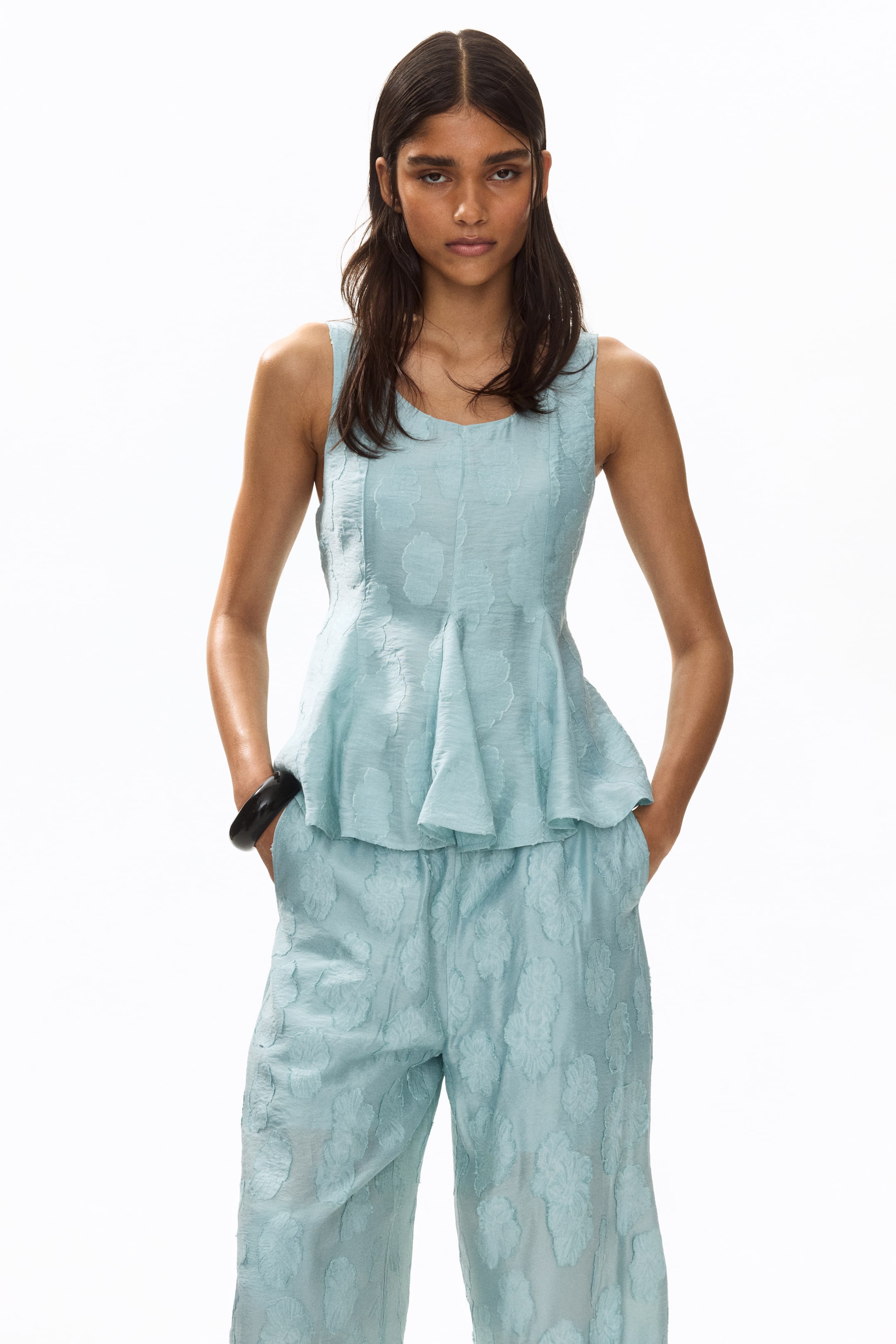 View larger image: Presenting a light blue sleeveless peplum top and matching relaxed-fit pants set, showcased from a frontal view. This ensemble features a subtle textured floral pattern throughout. The top is designed with a scoop neck and a flared peplum hem, offering a chic and elegant look.