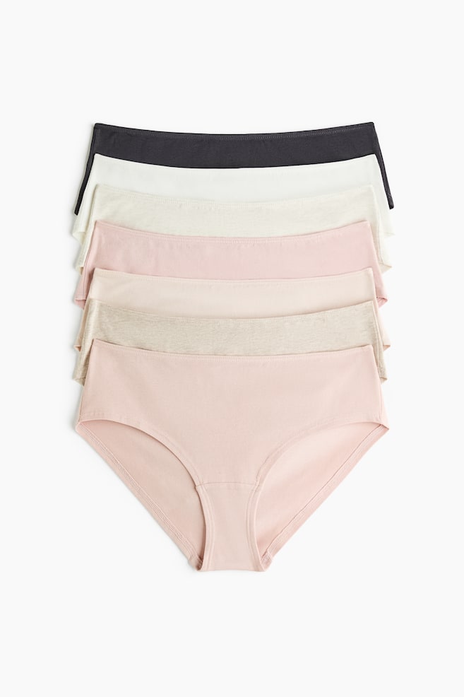 A seven-pack of mid-rise jersey briefs is laid flat in a vertical stack, showcasing a color palette of dark gray, white, cream, light pink, peach, heather gray, and dusty pink. The front-most dusty pink pair is fully visible from the front, featuring a classic brief cut with a stitched elastic waistband and a lined gusset. - 1