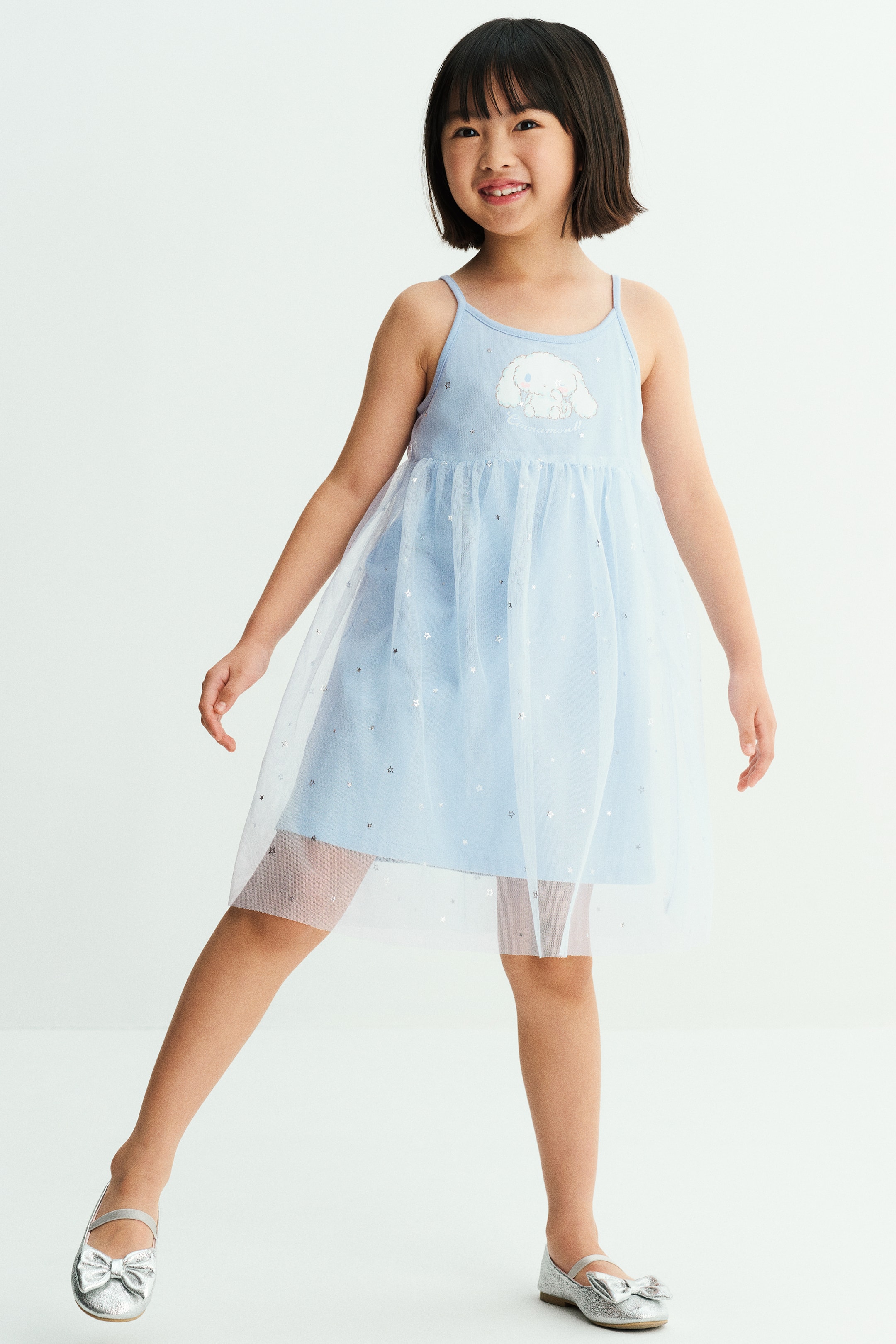 View larger image: A light blue jersey dress with narrow shoulder straps and a printed character on the chest is worn by the model. The dress features a flared skirt overlaid with a sheer, star-patterned tulle layer and is paired with silver metallic flat shoes.