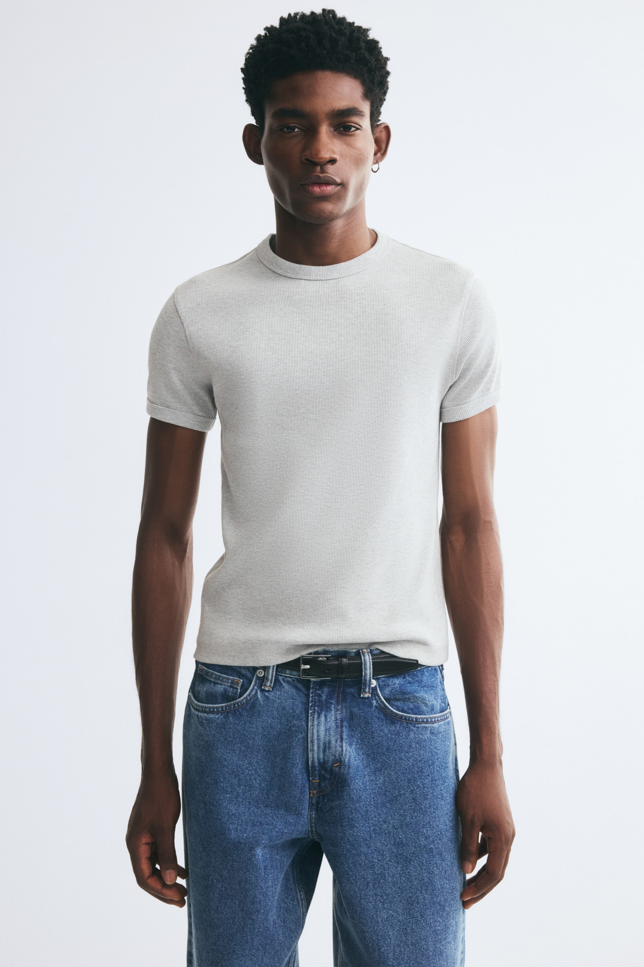 View larger image: The model is wearing a light grey ribbed cotton jersey T-shirt, featuring a slim fit, a round rib-trimmed neckline, and a straight-cut hem. The tee is styled untucked over blue denim jeans with a black belt.