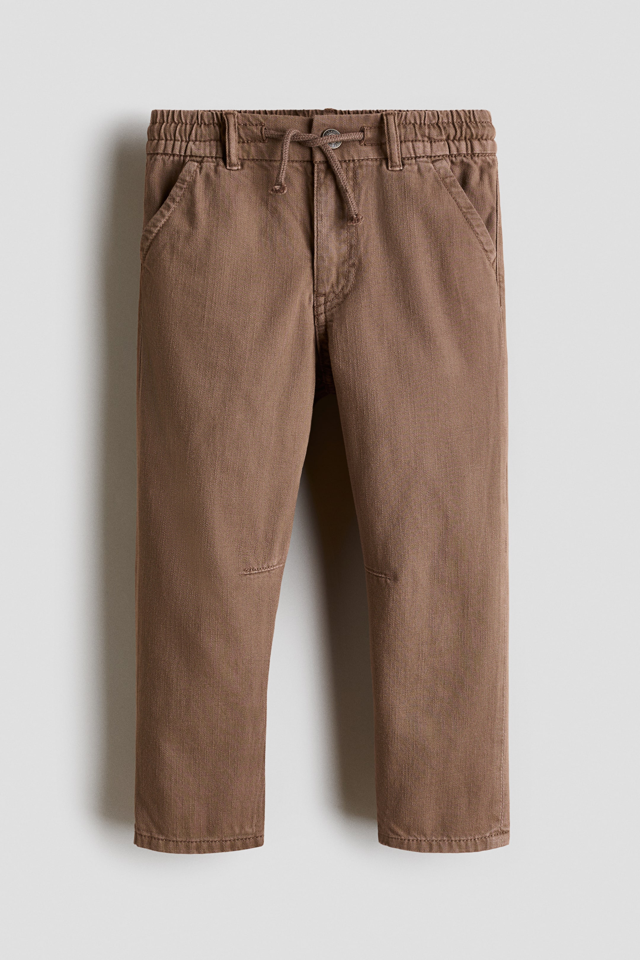 View larger image: Brown cotton twill trousers are shown from the front, featuring an elasticized drawstring waist and diagonal side pockets. They offer a relaxed fit through the hip and thigh with tapered legs and knee patches.