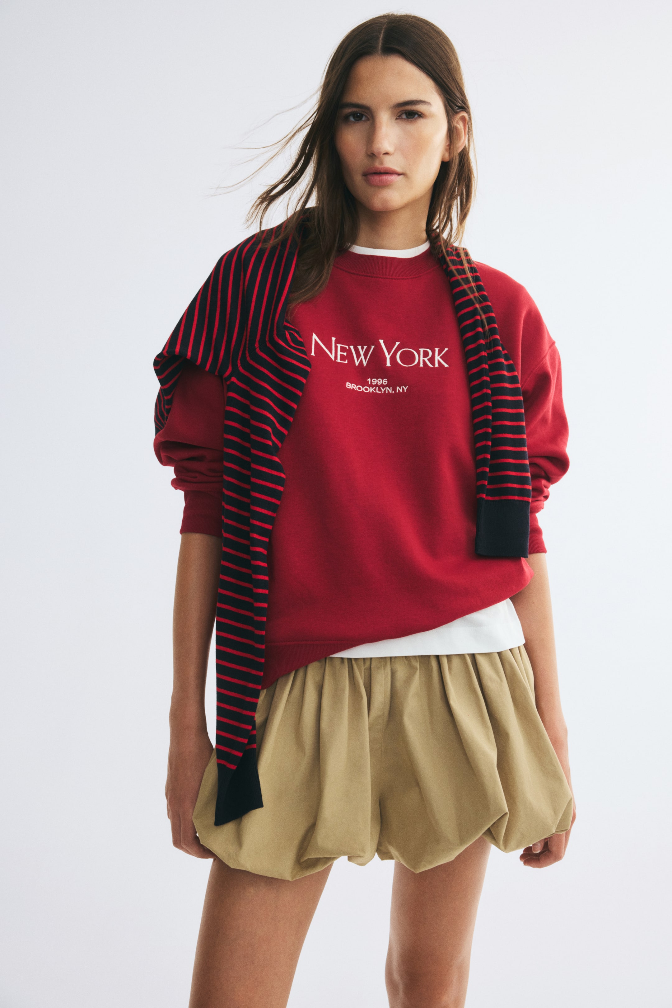 View larger image: A red oversized sweatshirt with a white "NEW YORK" text design on the front is being worn by the model. The sweatshirt features a ribbed crew neckline and dropped shoulders, and is styled with a black and red striped sweater draped over the shoulders and light beige bubble shorts.