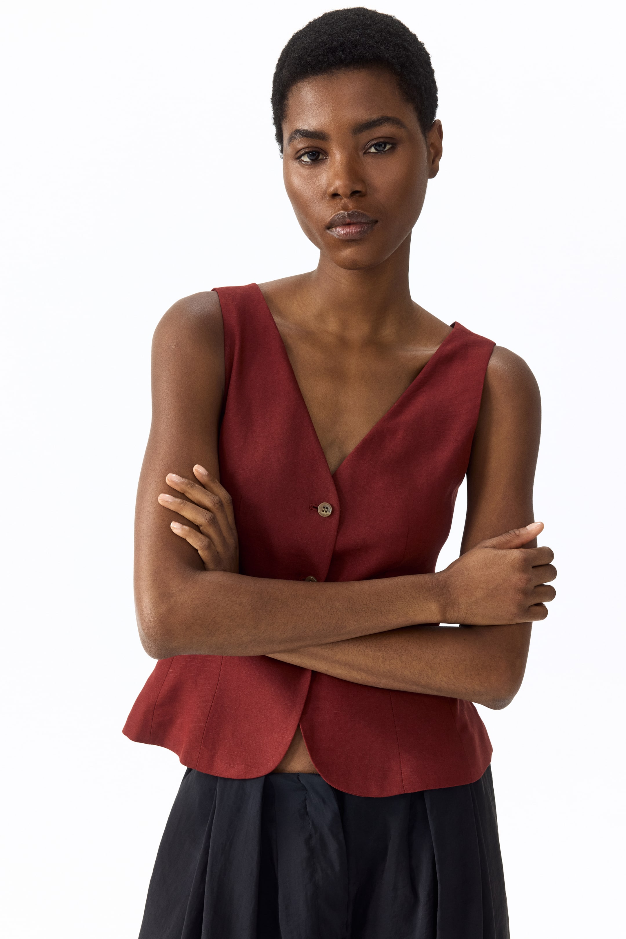 查看大圖: 這位模特穿著一件量身訂製的深紅色馬甲，採用織布製成，前面設計有V領口和按鈕扣合。模特雙臂交叉於馬甲上，搭配深色長褲，展現出時尚的風格。