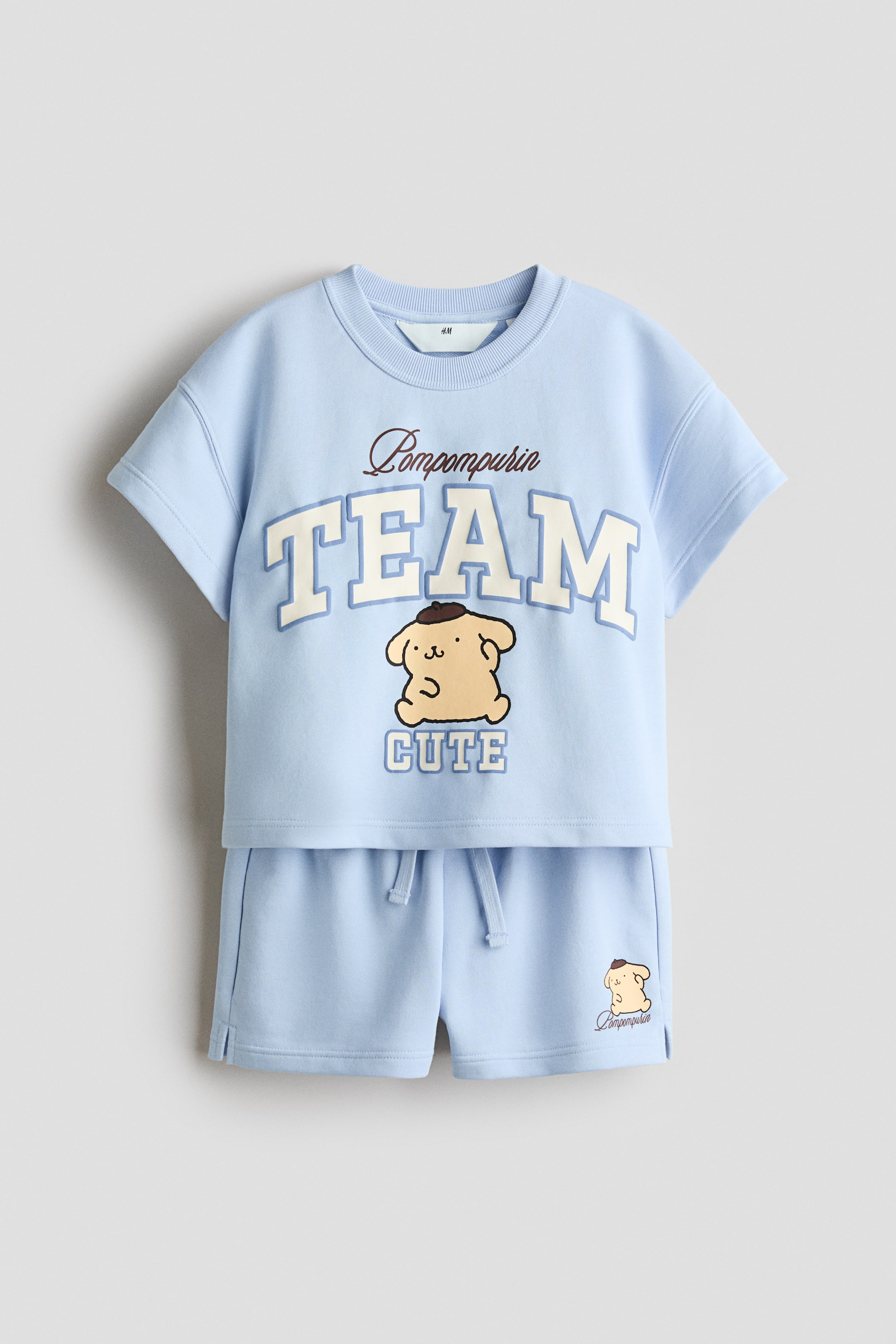 拡大画像を表示: ライトブルーの2ピースセットは、プリントされたスウェット生地のTシャツとショーツで構成されており、正面からのビューが示されています。Tシャツは、リブトリム付きのネックライン、ドロップショルダー、「Pompompurin TEAM CUTE」というテキストとポムポムプリンのキャラクターグラフィックを前面に展示しています。一方、コーディネートされたショーツは、伸縮性のあるドローストリングウエストと左脚に小さなポムポムプリンのキャラクタープリントを特徴としています。