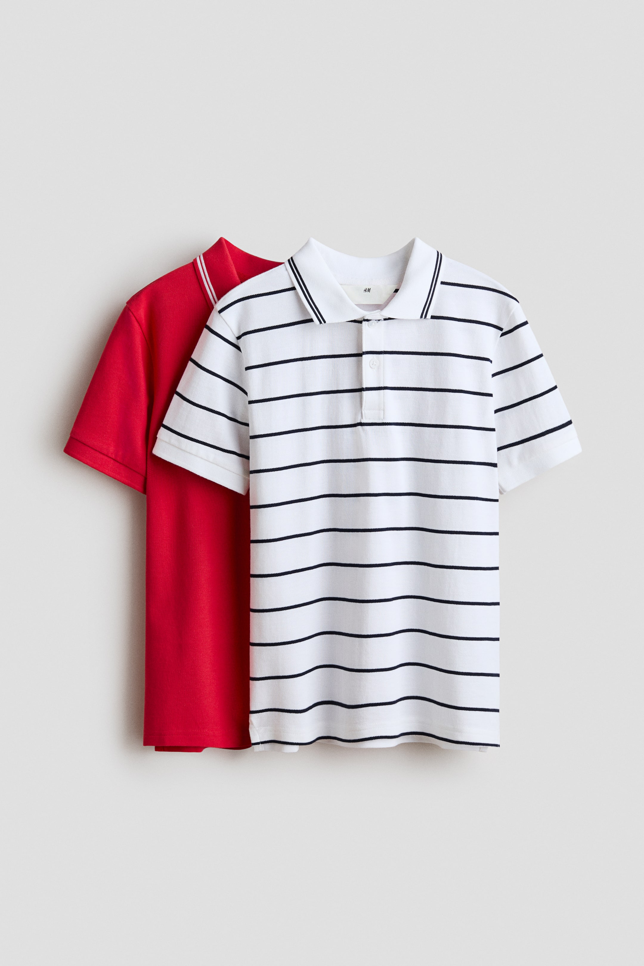 Grösseres Bild ansehen: Zwei Poloshirts sind ausgestellt, übereinander gelegt, beide nach vorne gerichtet. Das vordere Poloshirt ist weiss mit horizontalen marineblauen Streifen, verfügt über einen weissen Kragen mit dünner marineblauer Kante, eine Zwei-Knopf-Leiste und gerippte kurze Ärmel, während ein einfarbiges rotes Poloshirt mit weisser Kante am Kragen teilweise dahinter sichtbar ist.