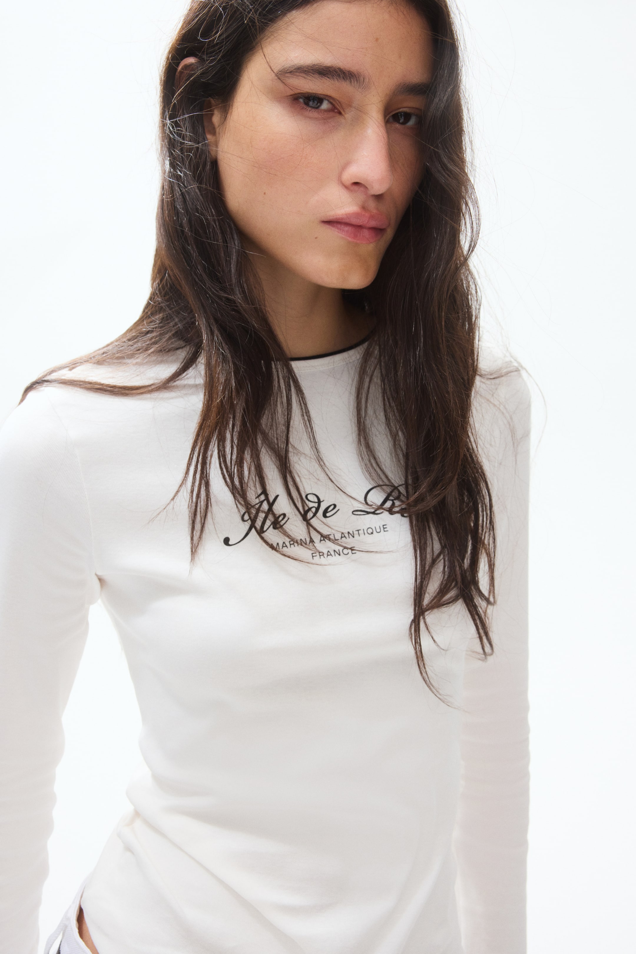 View larger image: The model is wearing a white long-sleeved top, viewed from the front with a slight right angle. It features a round neckline with a thin black trim. Across the chest, there's black script text reading "Ile de Ré MARINA ATLANTIQUE FRANCE," partially obscured by hair.