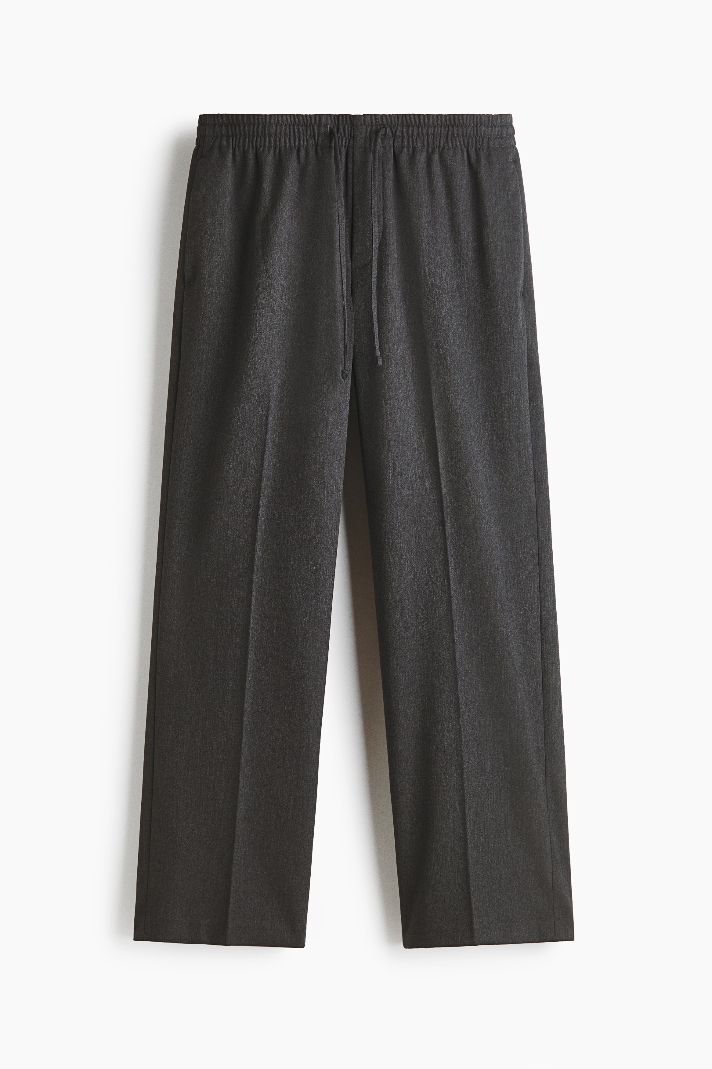 Tailored joggers in a dark, woven fabric are worn by the model, featuring a relaxed fit, an elasticated drawstring waist, and visible creases down the front of the legs. The joggers are styled with a cream-coloured polo shirt, a black leather jacket with a brown shearling collar, and black leather shoes.