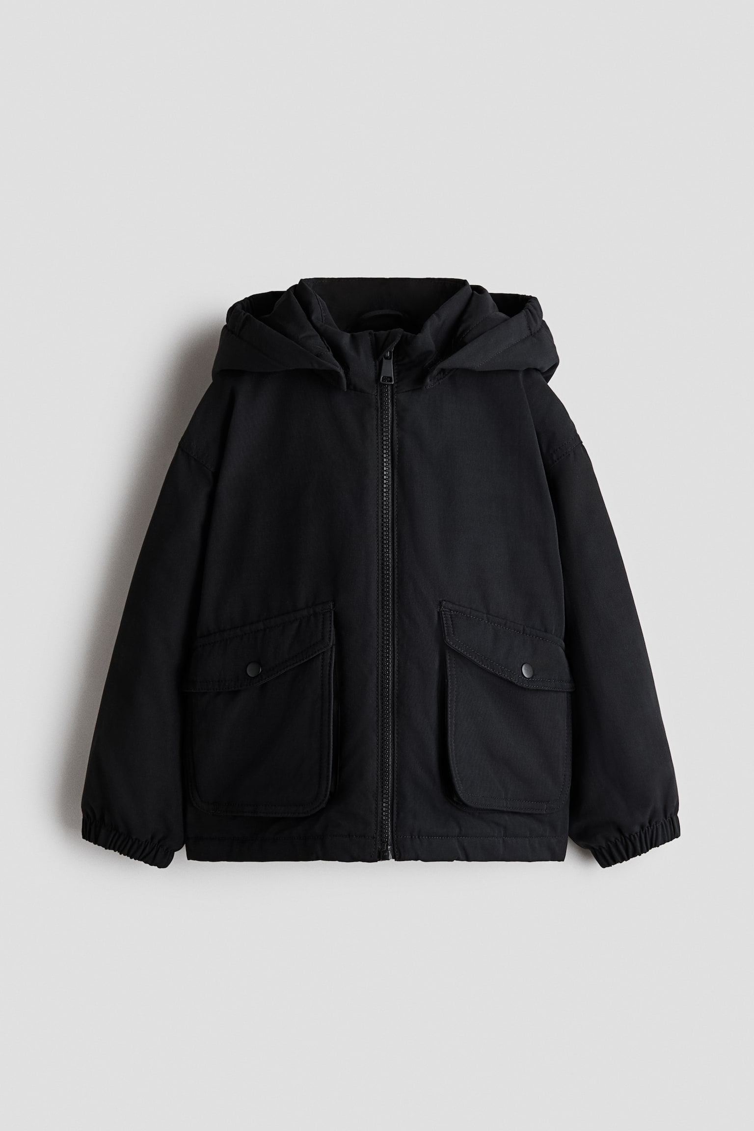 A black, lightly padded jacket is shown from the front, featuring a full-length zip, an attached hood, and dropped shoulders with elasticated cuffs. Two prominent bellow pockets with flap closures are positioned on the lower front.