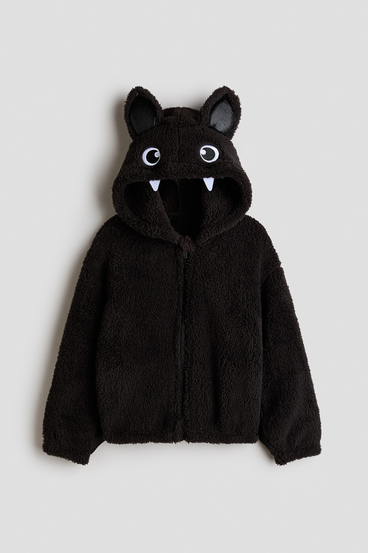 A black teddy fleece jacket is facing forward. Its hood is detailed with pointed ears, white eyes with black pupils, and two white fangs. A full-zip closure is running down the center front. - 1