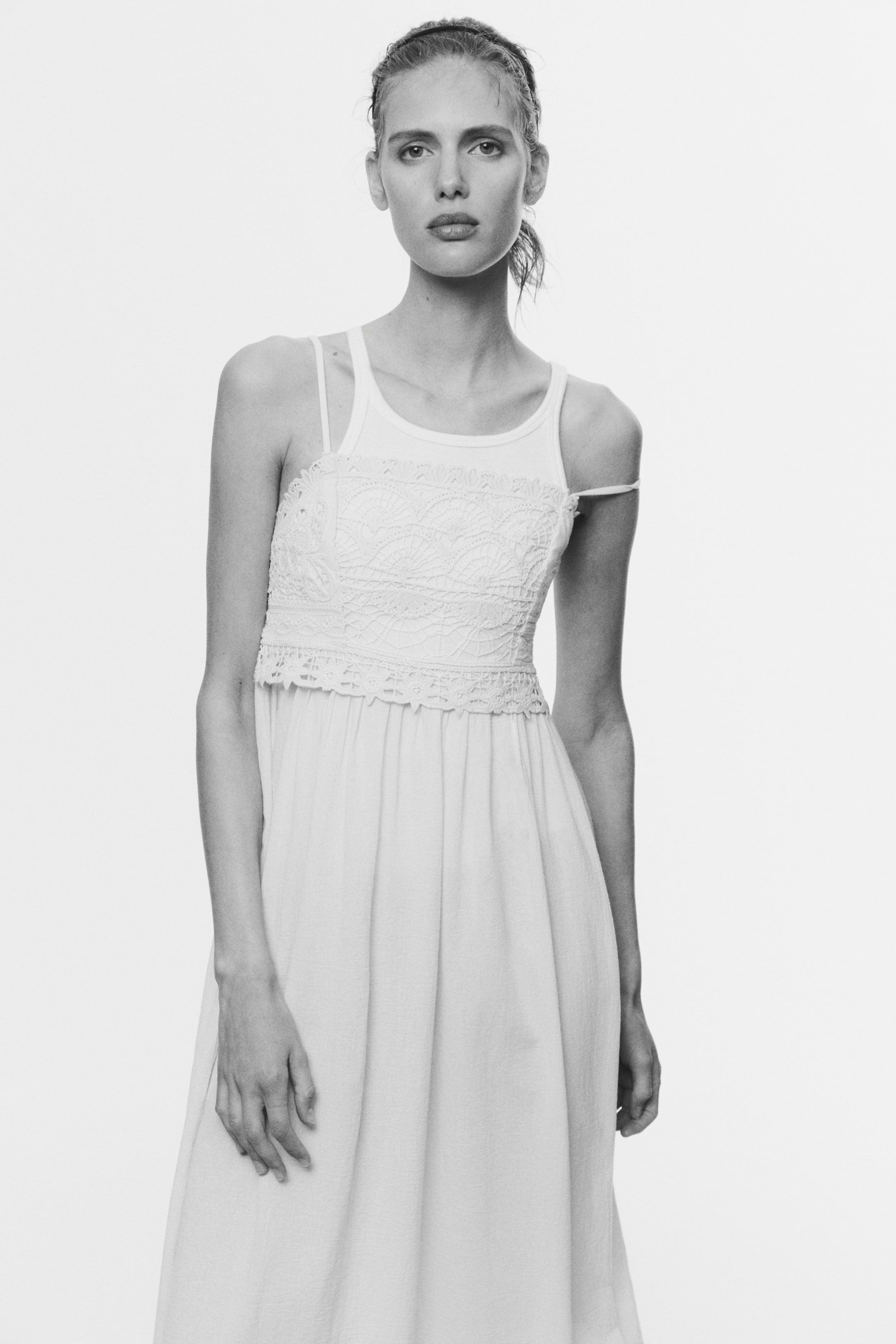 View larger image: A light-hued dress showcases a camisole-style top with slender shoulder straps and an intricately designed crochet panel across the bust. The dress gracefully transitions into a loose skirt beneath a decorative trim at the empire waist, with one strap elegantly slipping down the right arm.