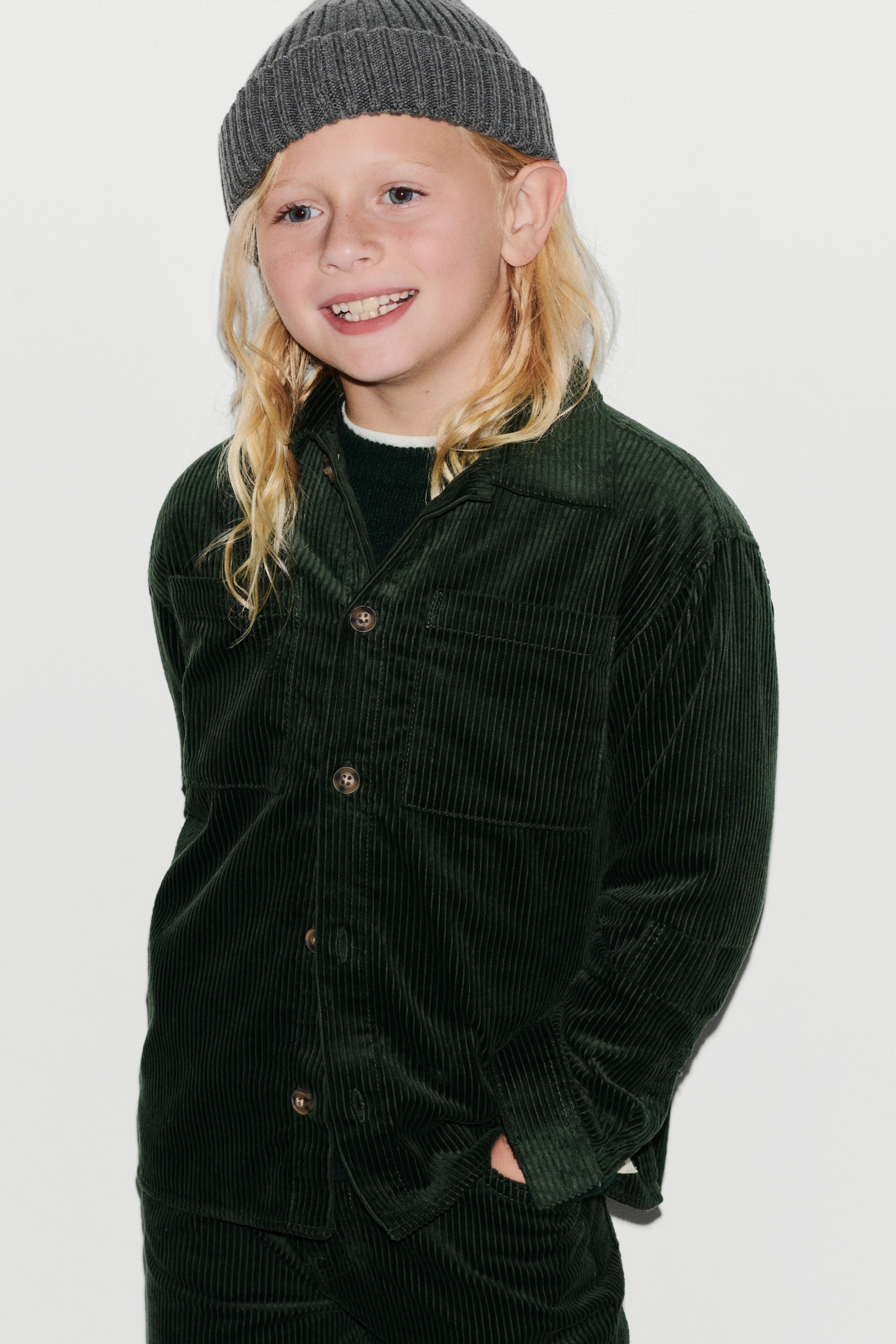 View larger image: A dark green corduroy overshirt is shown from the front, featuring a classic collar, button fastening, and two chest patch pockets. The loose-fit shirt is layered over a dark crew neck jumper, displaying its soft ribbed texture and relaxed fit.