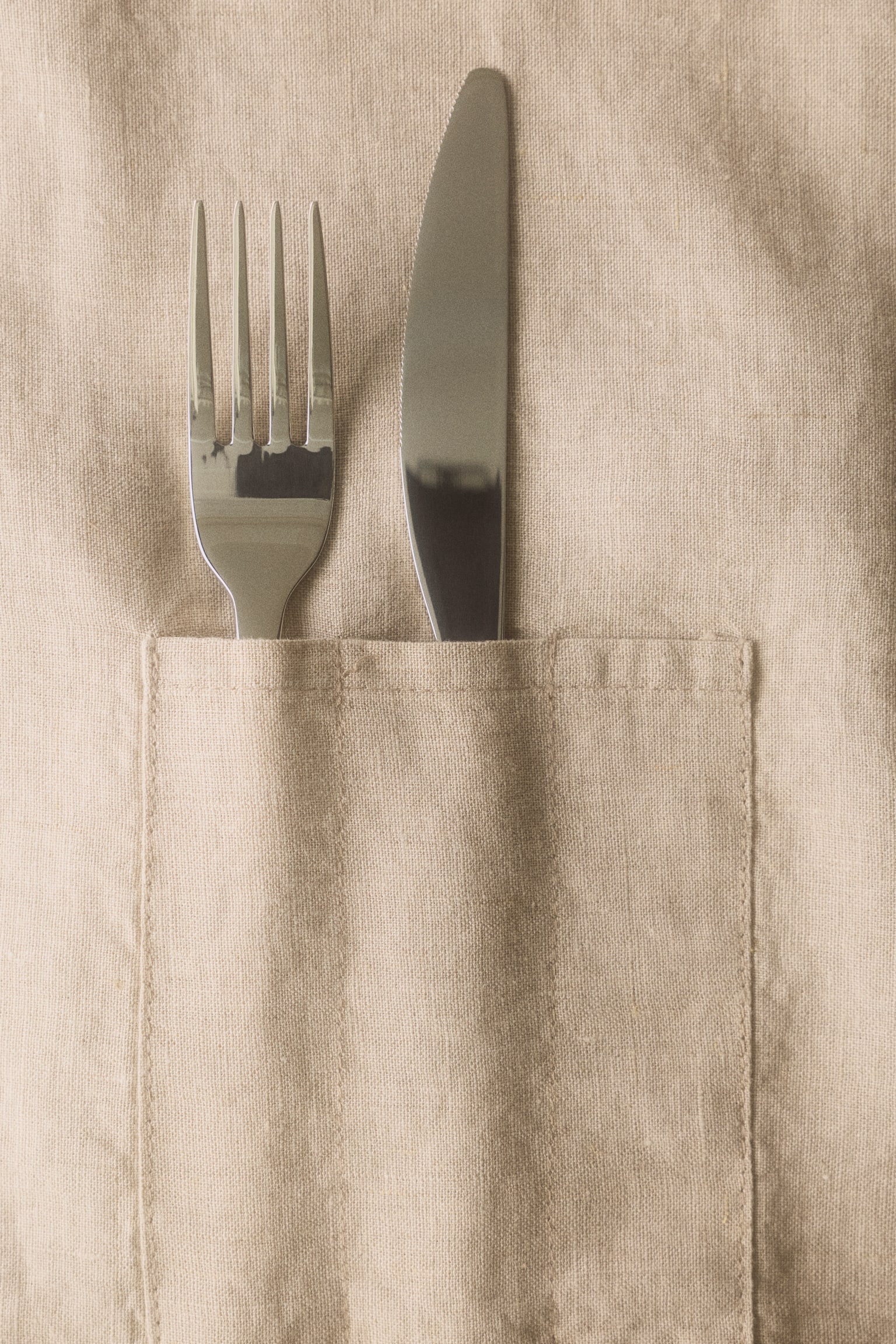 A light beige linen weave cutlery pouch is displayed front-facing, featuring a fold-over design. It contains a spoon, fork, and knife within its separate compartments. - 1