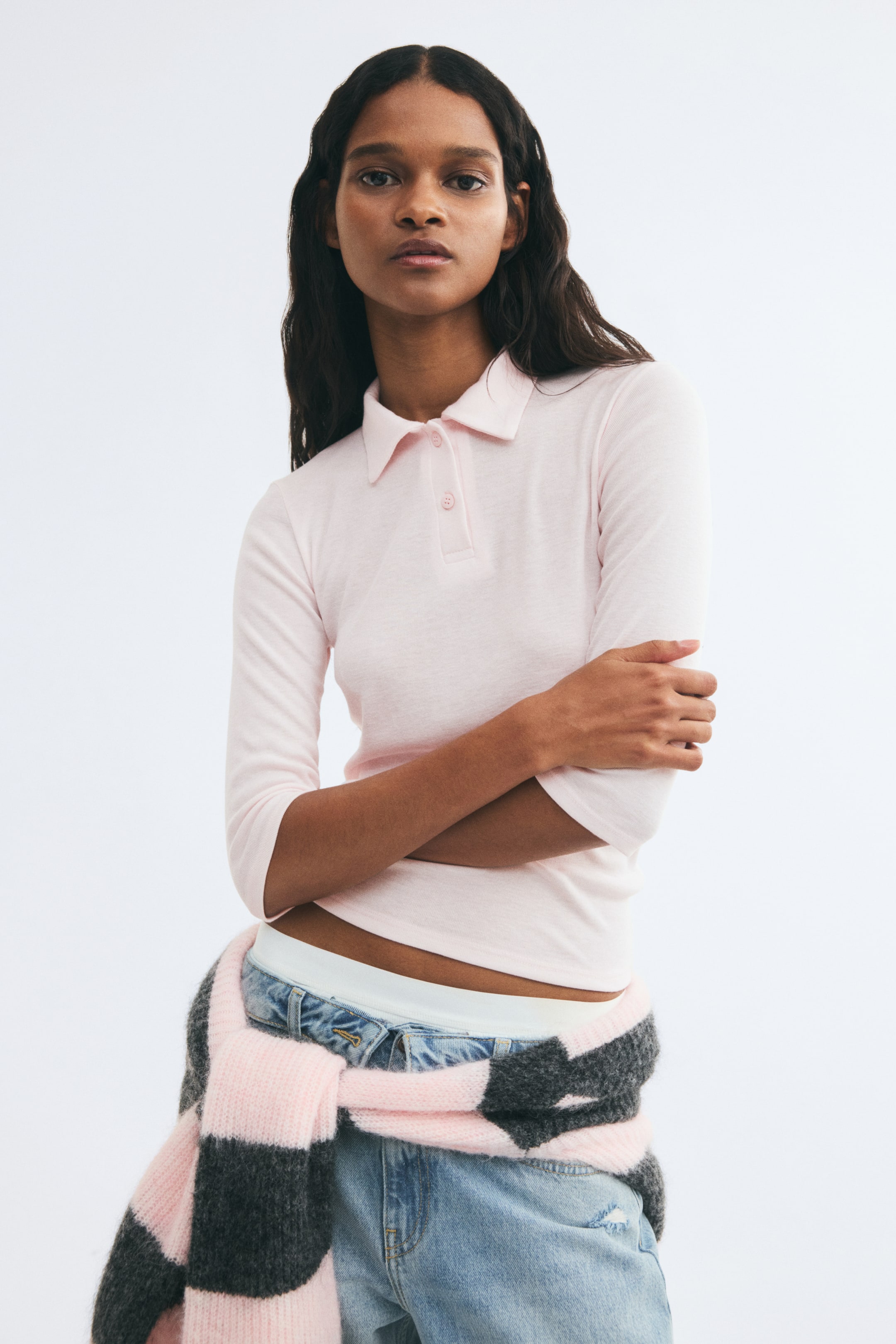 Grotere afbeelding bekijken: Het model draagt een lichtroze poloshirt van fijne ribjersey, voorzien van een kraag, een knoopsluiting, 3/4-mouwen en een rechte zoom. Het shirt is gestyled met denim jeans in een lichte wassing en een gebreide trui met roze en grijze strepen, die om de taille is geknoopt.