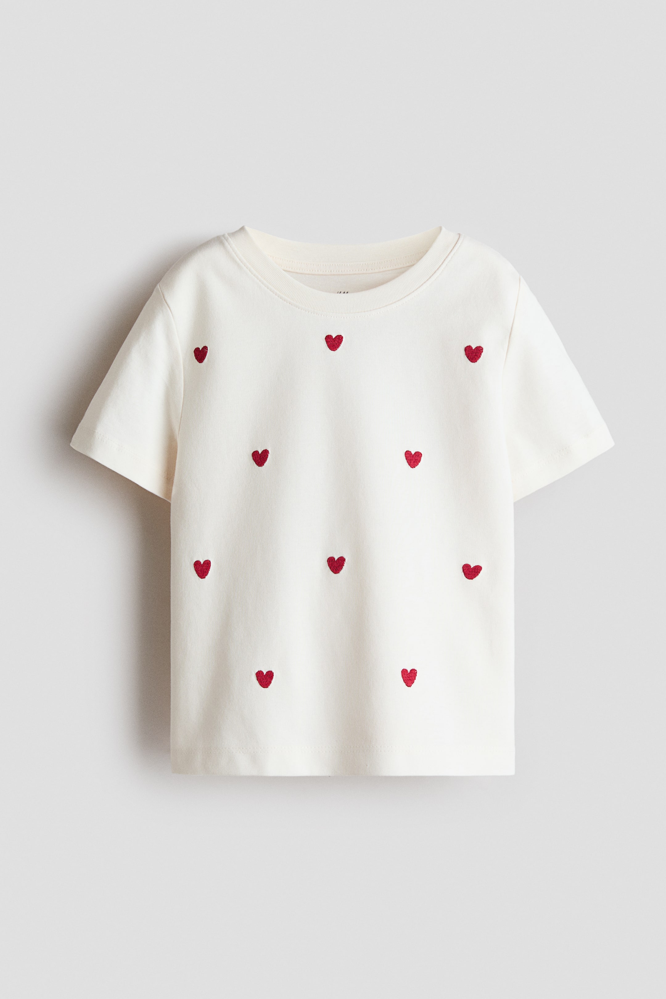Vezi imaginea mai mare: Un tricou crem din jerseu de bumbac moale este orientat spre față, având un decolteu rotund cu margini ribate și mâneci scurte. Motive mici de inimă roșie sunt împrăștiate pe partea din față.