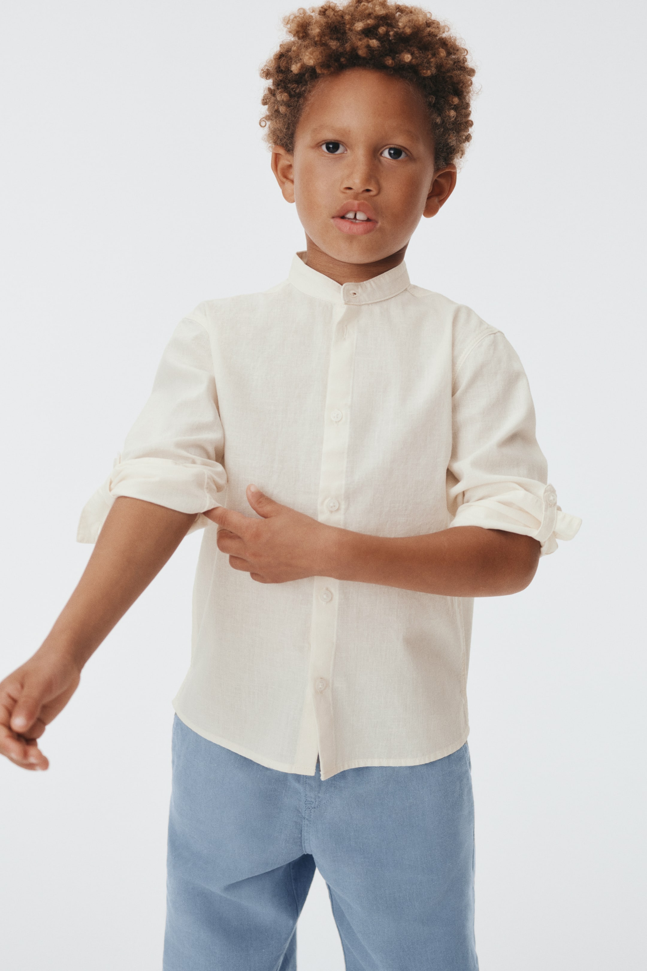 הצגת תמונה גדולה יותר: חולצה ארוכת שרוול בצבע קרם עם טקסטורה מוצגת מלפנים, עם צווארון מנדרין, כפתרה מלאה ושרוולים מגולגלים ומאובטחים בלשוניות וכפתורים, מעוצבת מעל מכנסיים בצבע כחול בהיר.