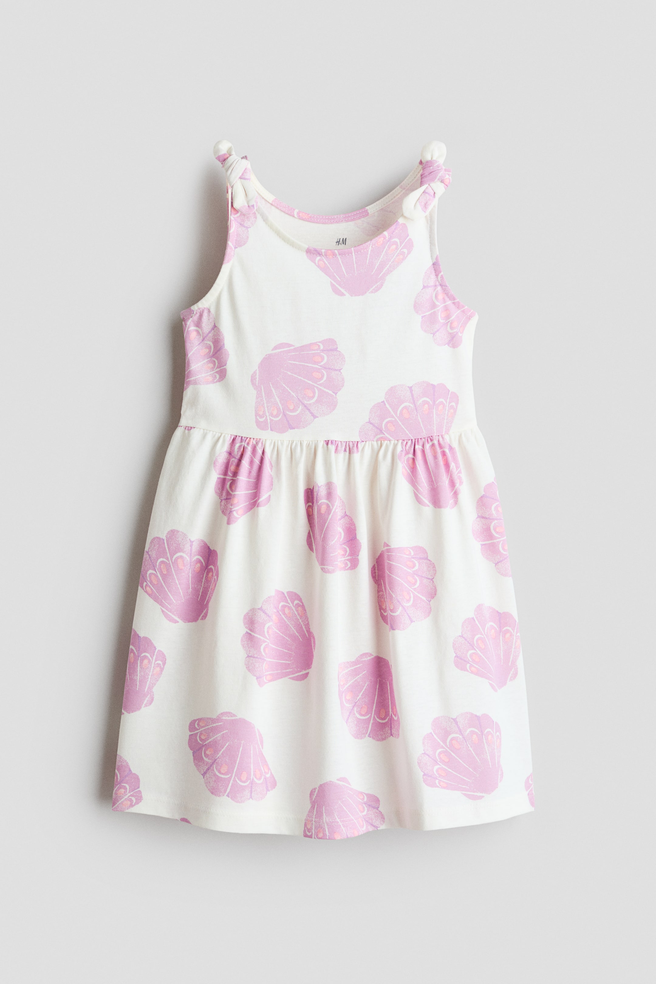 View larger image: The dress showcases a white base adorned with an all-over pink seashell print, displayed from the front. It is crafted with a rounded neckline and shoulder straps that are tied into knots. The waistline is gathered, and the skirt flares out slightly.