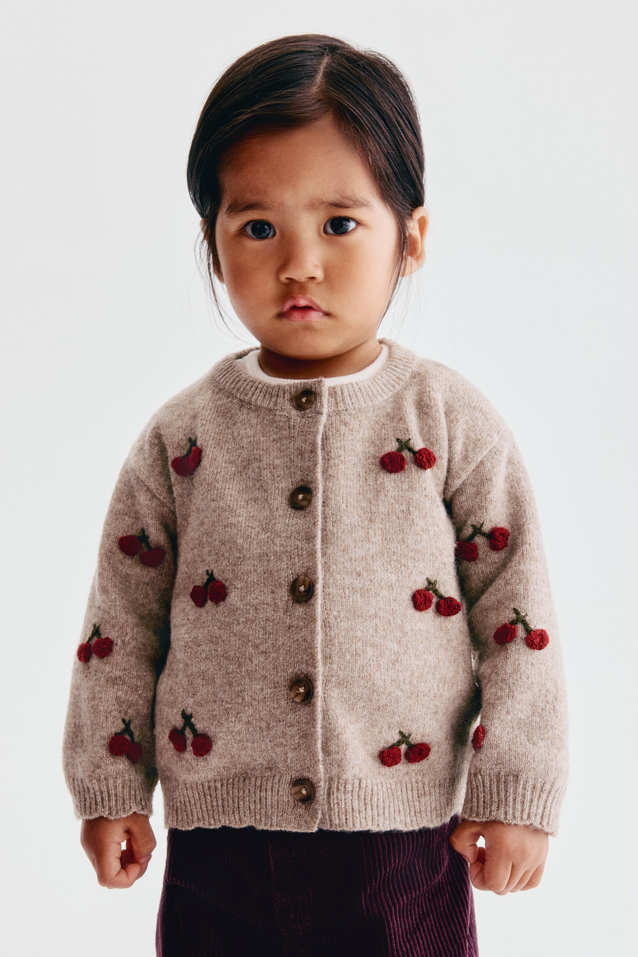 Agrandir l'image: Un cardigan en maille beige est présenté de face, avec un motif intégral de cerises rouges brodées à tiges vertes. Le cardigan affiche une fermeture boutonnée sur le devant avec cinq boutons bruns, un col rond côtelé et des finitions côtelées aux poignets et à l’ourlet.