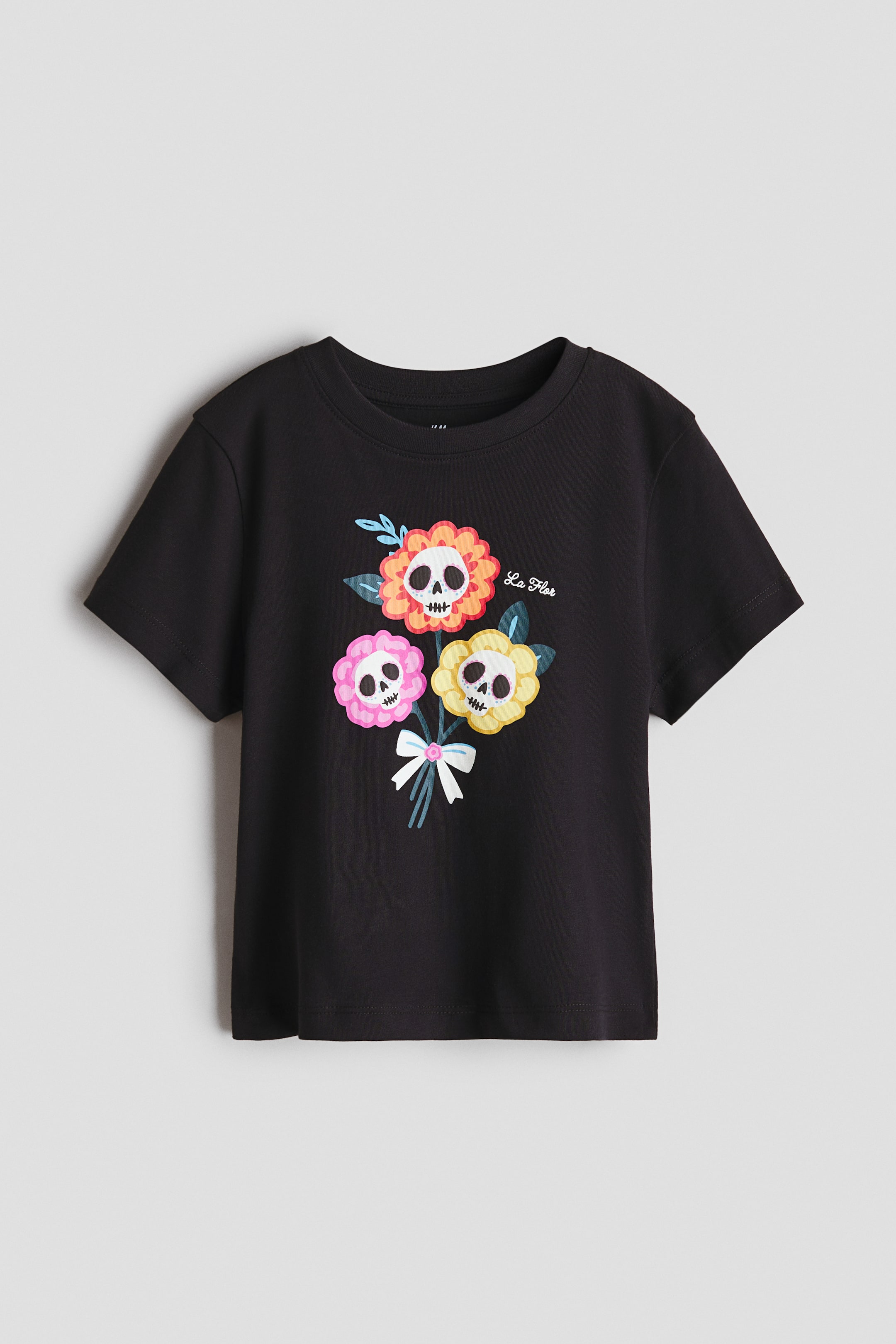 View larger image: A black T-shirt showcases a front-facing graphic print. The print depicts a bouquet of three flowers with skull faces in their centers—one orange, one pink, and one yellow—tied together with a light blue and white bow, with the text "La Flor" appearing near the orange flower.