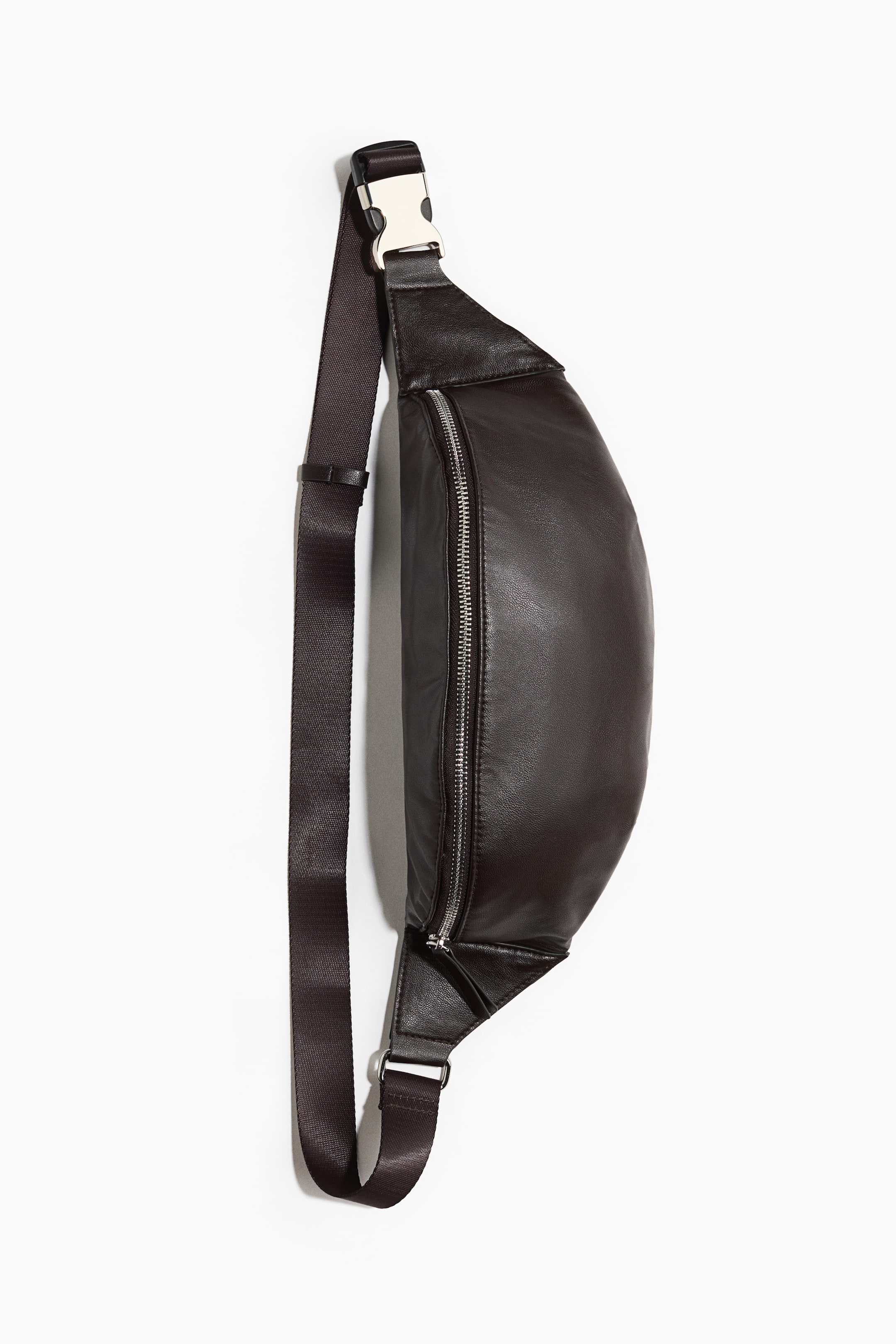 View larger image: A dark brown crossbody bag in coated fabric is hanging vertically from a front angle, showcasing its curved, crescent-shaped main compartment. The bag features a silver metal zipper along the top opening and an adjustable dark brown woven strap with a silver metal snap buckle.