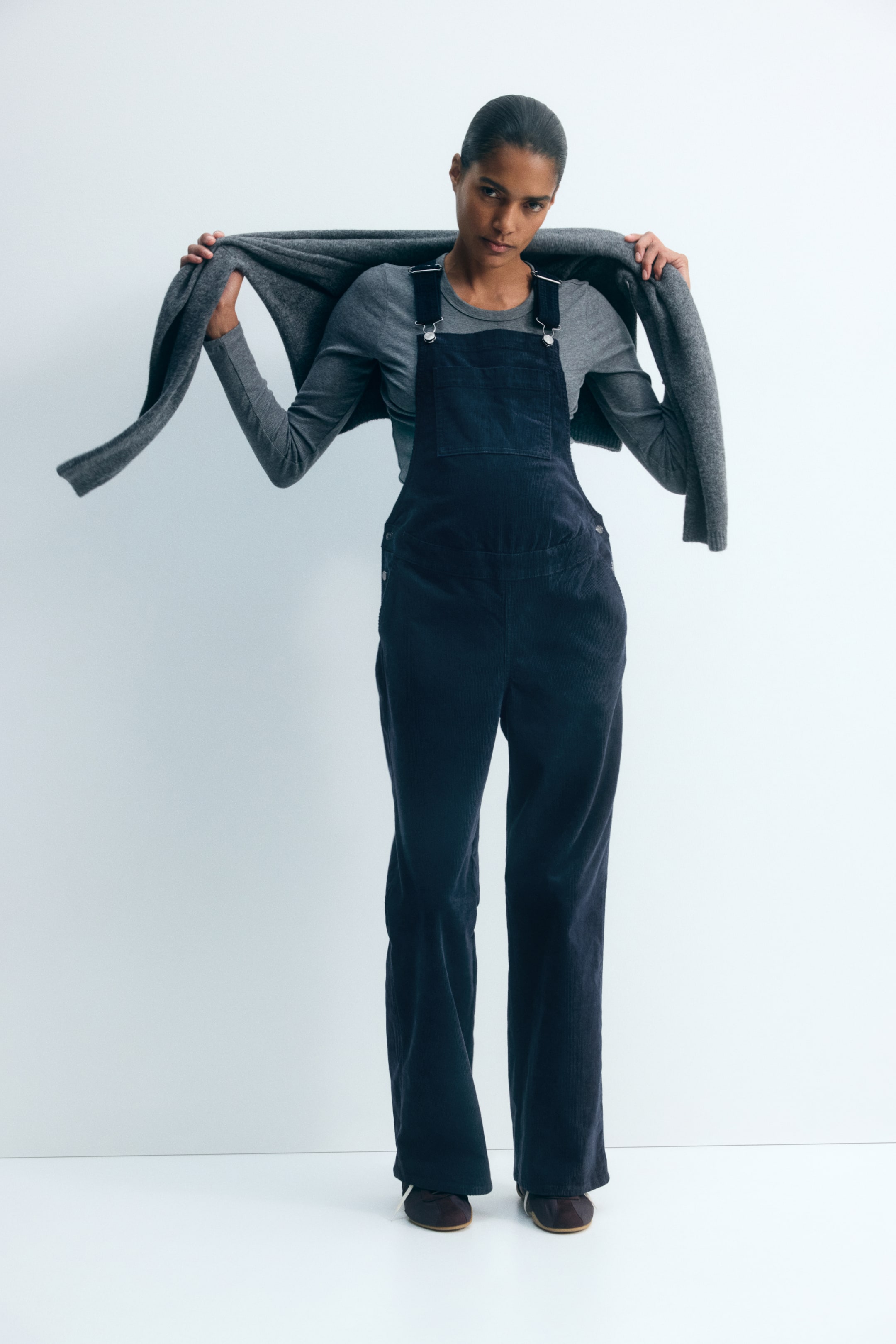 View larger image: Dark navy cotton corduroy dungarees featuring a square neckline, adjustable shoulder straps, and a front patch pocket are worn by the model over a grey long-sleeved top. The model is holding a grey knit sweater draped over their shoulders.